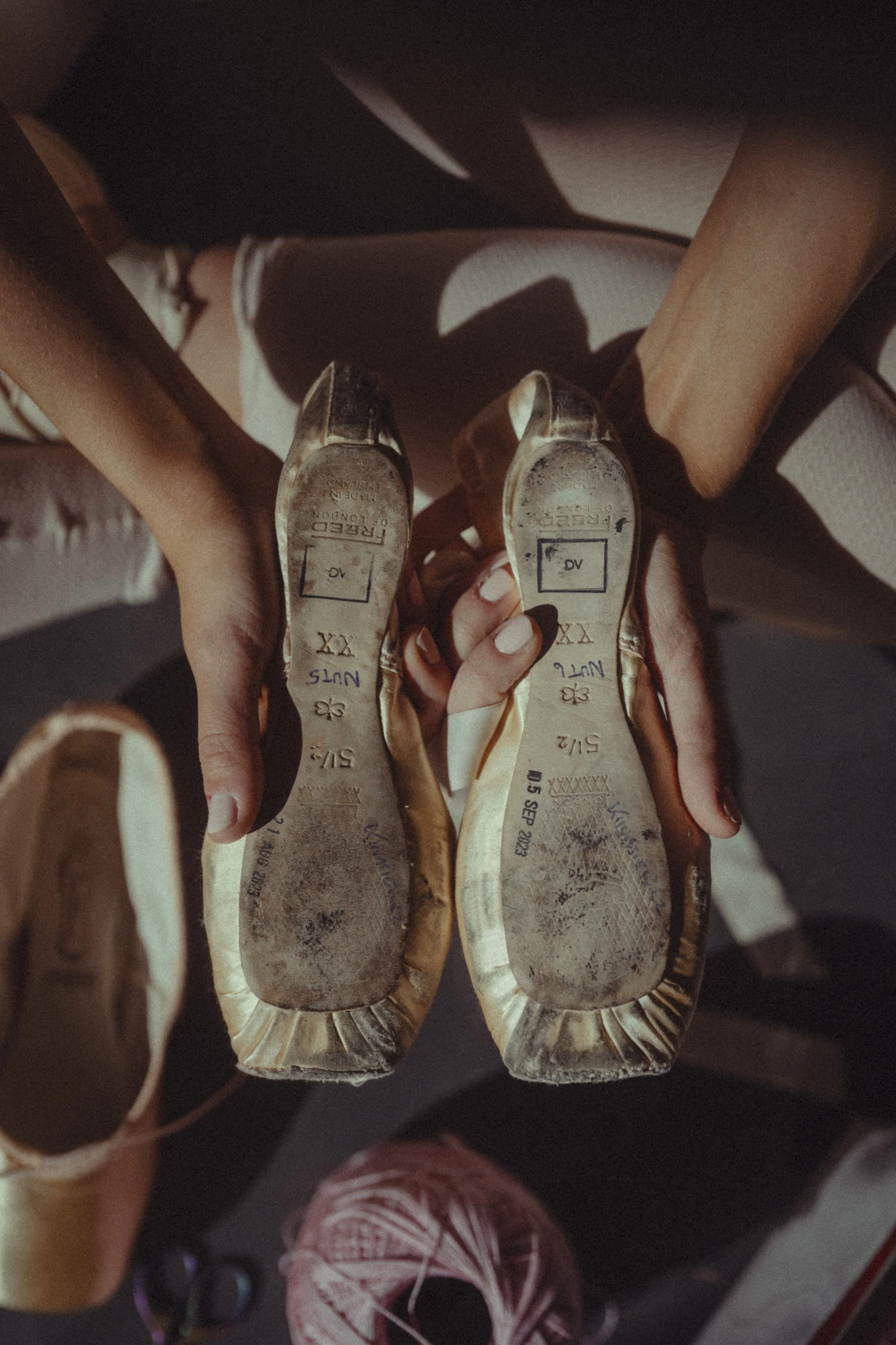 A person holding a pair of worn gold-colored high heel shoes, showing the soles marked with size and other details, with sunlight casting shadows on the shoes and surroundings.