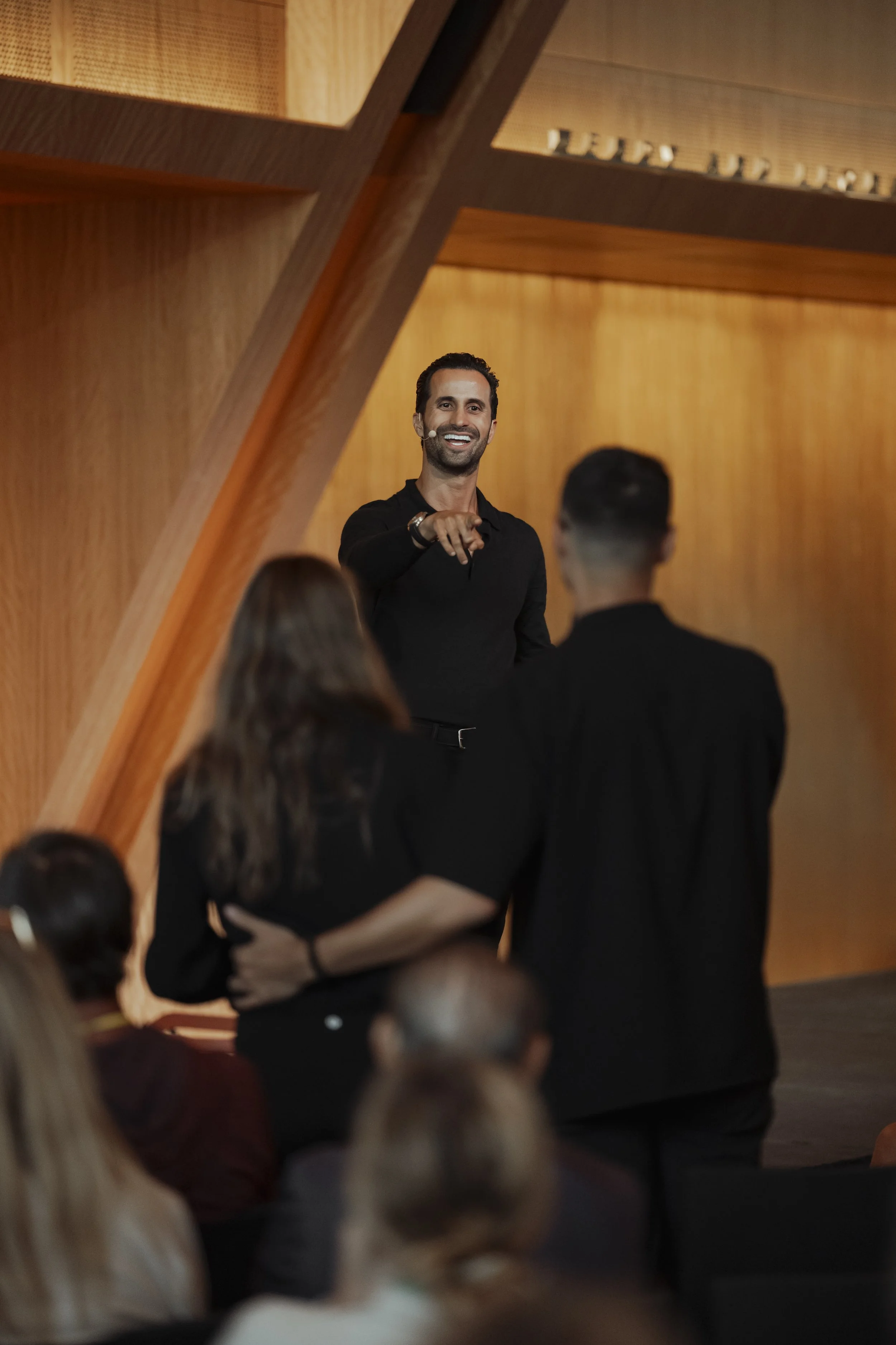 A man with dark hair and a beard, wearing a black shirt, standing on a stage, smiling and pointing towards the audience, with a wooden background and audience members in the foreground.