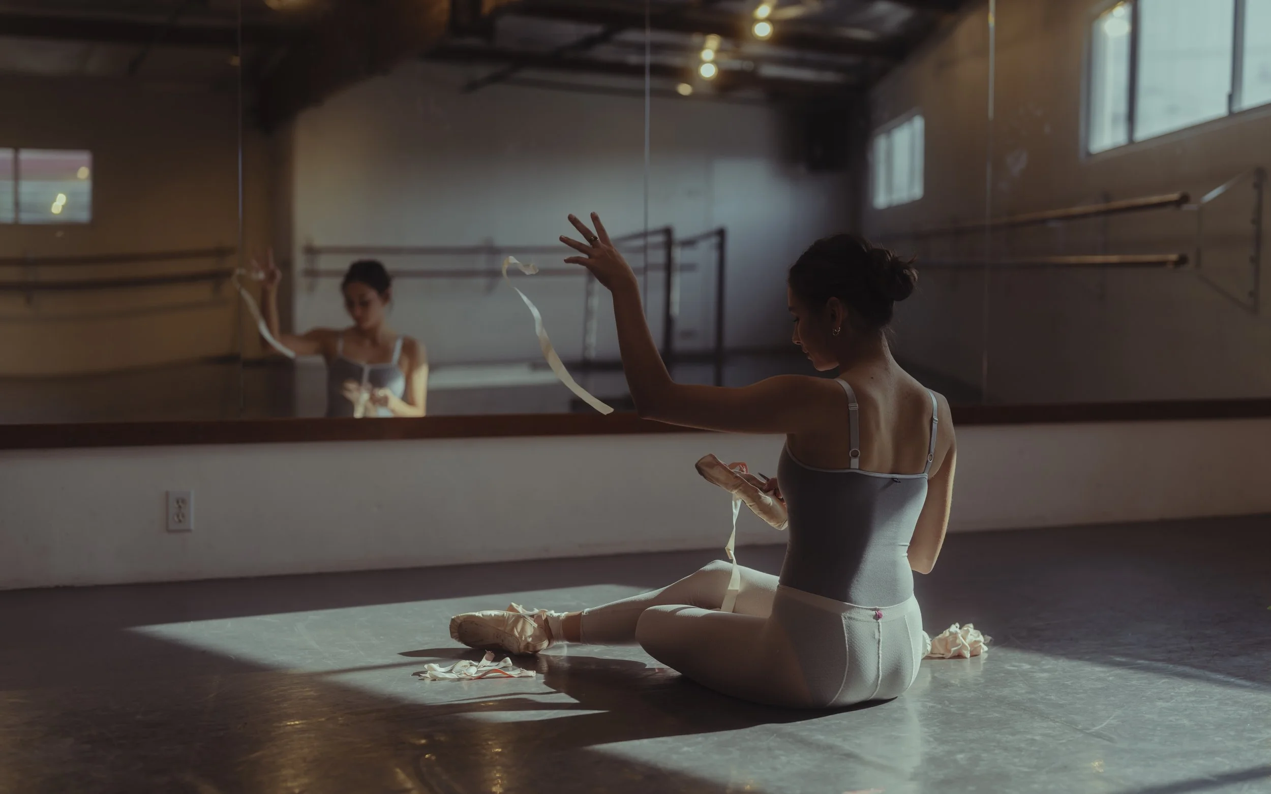 Ballet dancer sitting on the floor of a dance studio with ribbons, wearing a leotard and tights, looking at her phone, reflected in the mirror.