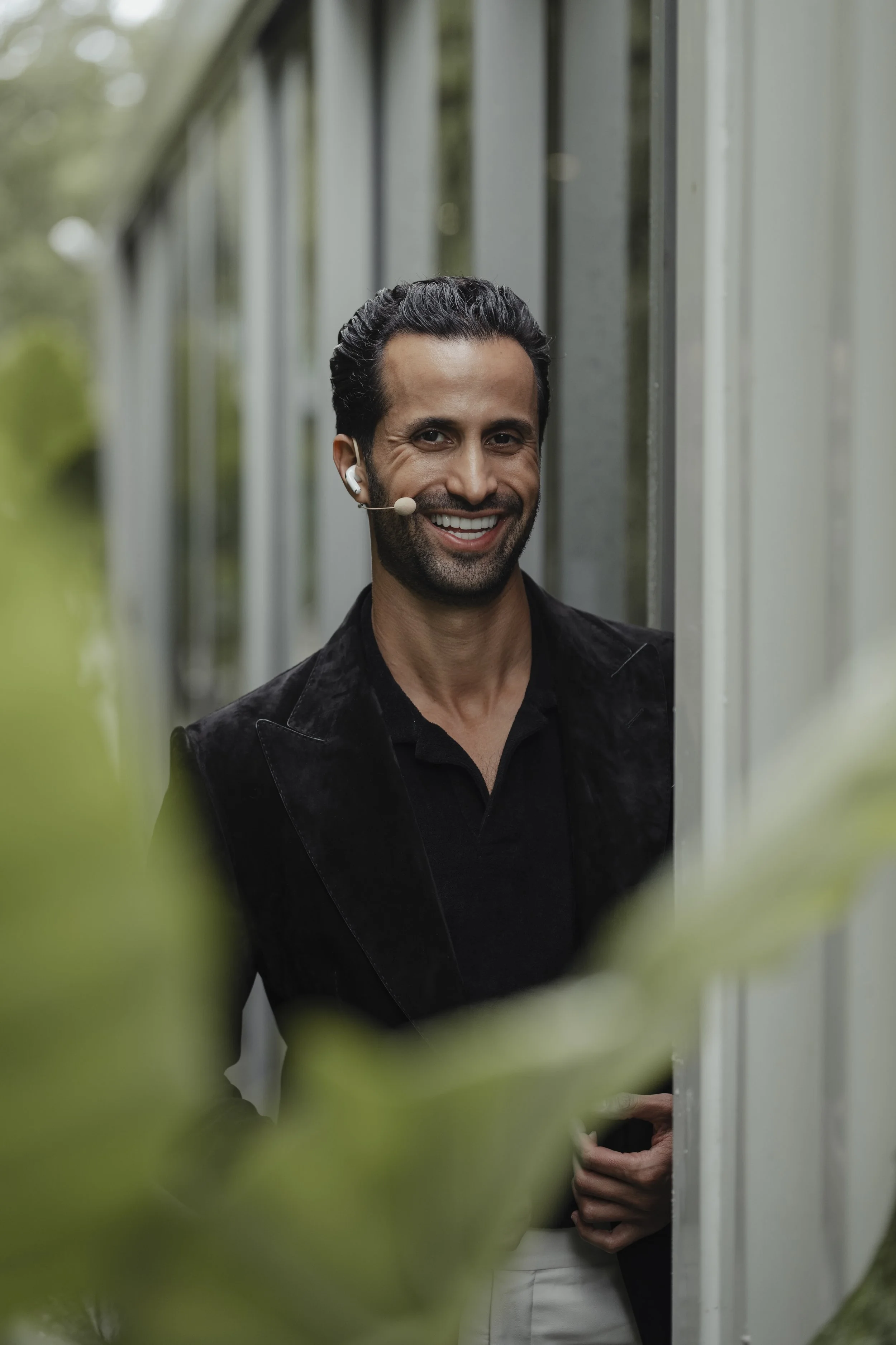 A man with dark hair, beard, and a headset smiling while standing outdoors near a building with large windows, surrounded by greenery.