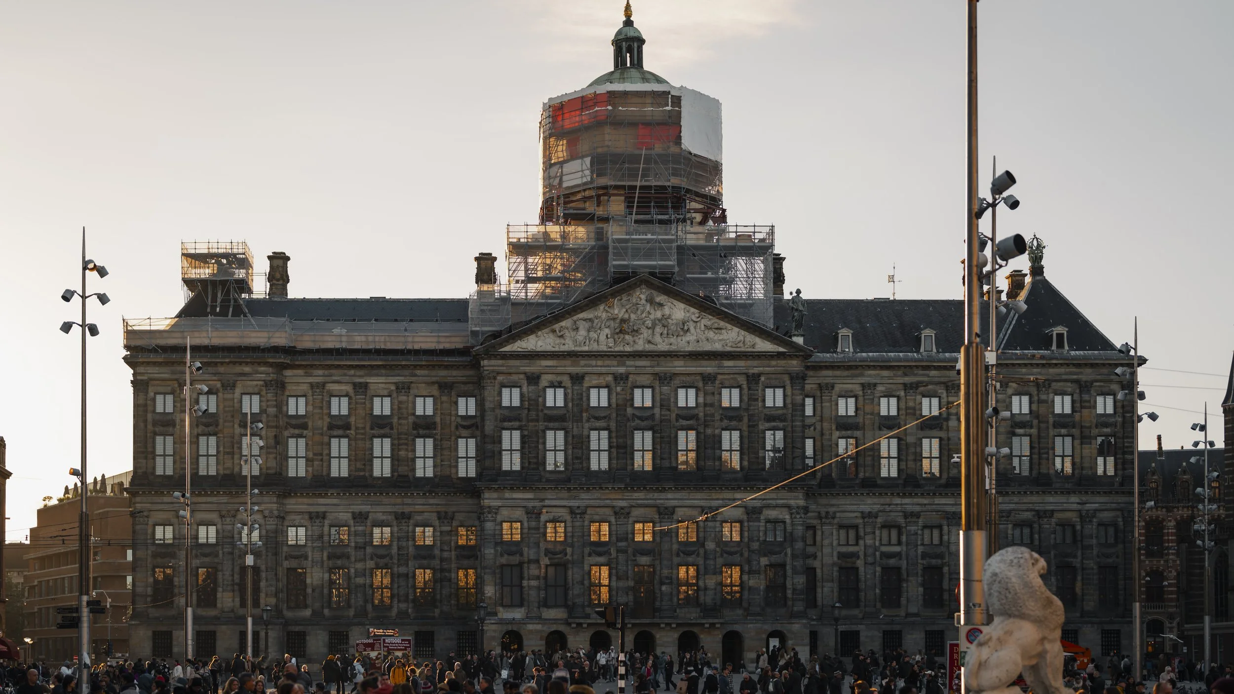 A large historic building with scaffolding around the upper part, possibly under renovation, during sunset. The building has many windows, some reflecting sunlight, and features statues on the roof. In the foreground, a crowded public square with man