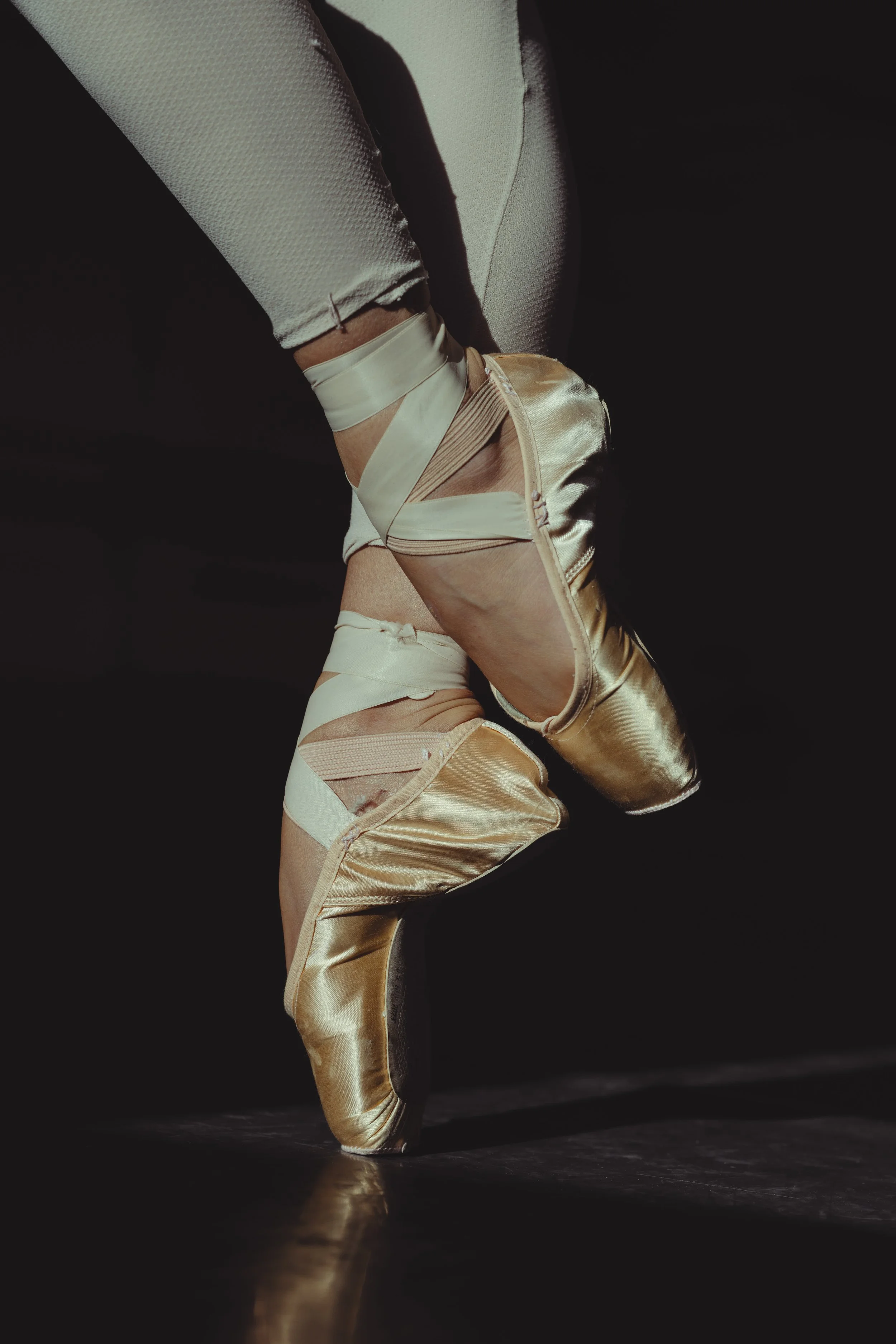 Close-up of a ballet dancer's feet in gold satin pointe shoes, with a reflection on the dark floor.