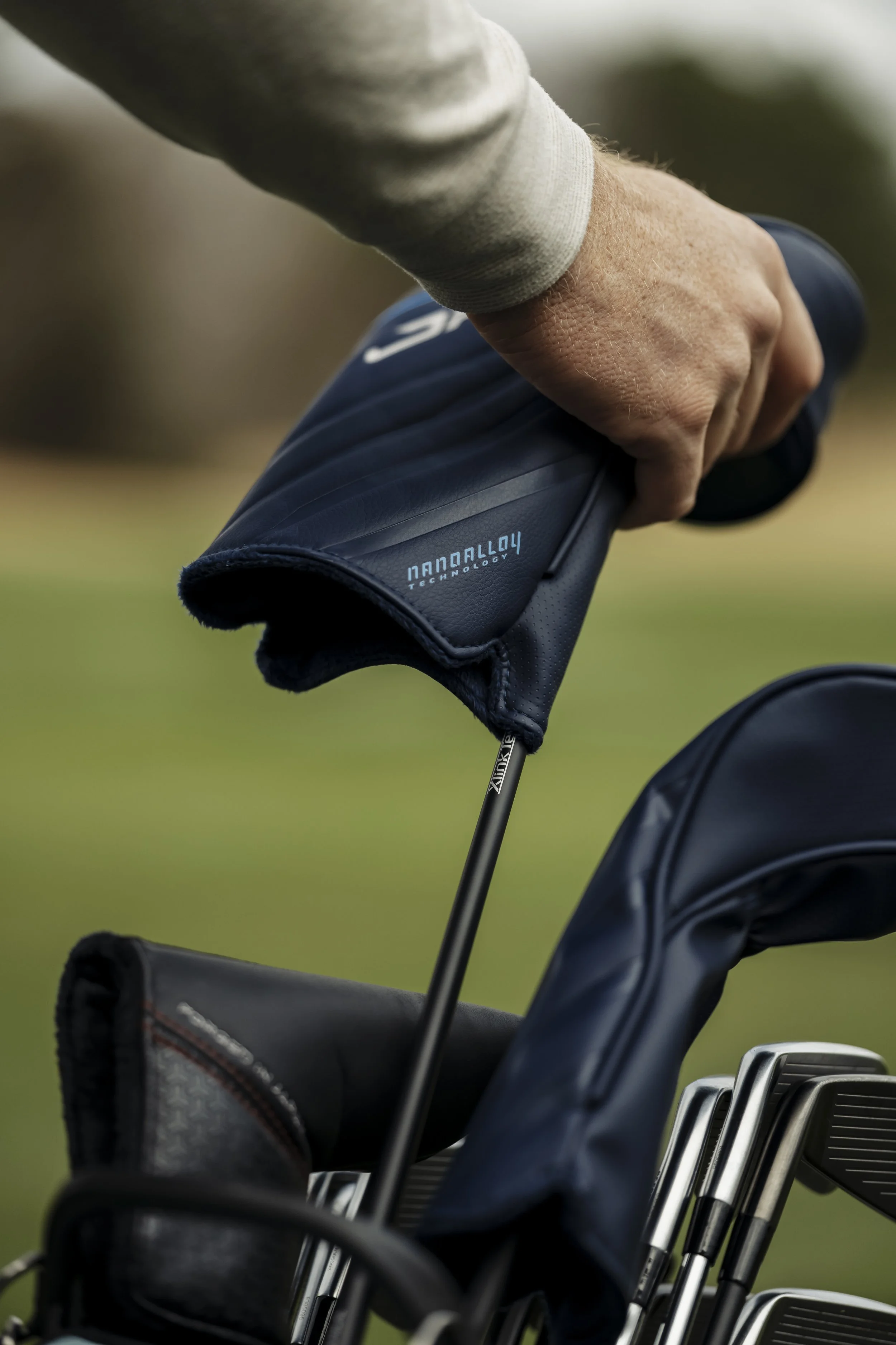 A person holding a golf club with a headcover, near a golf bag with additional clubs, on a green outdoor golf course.