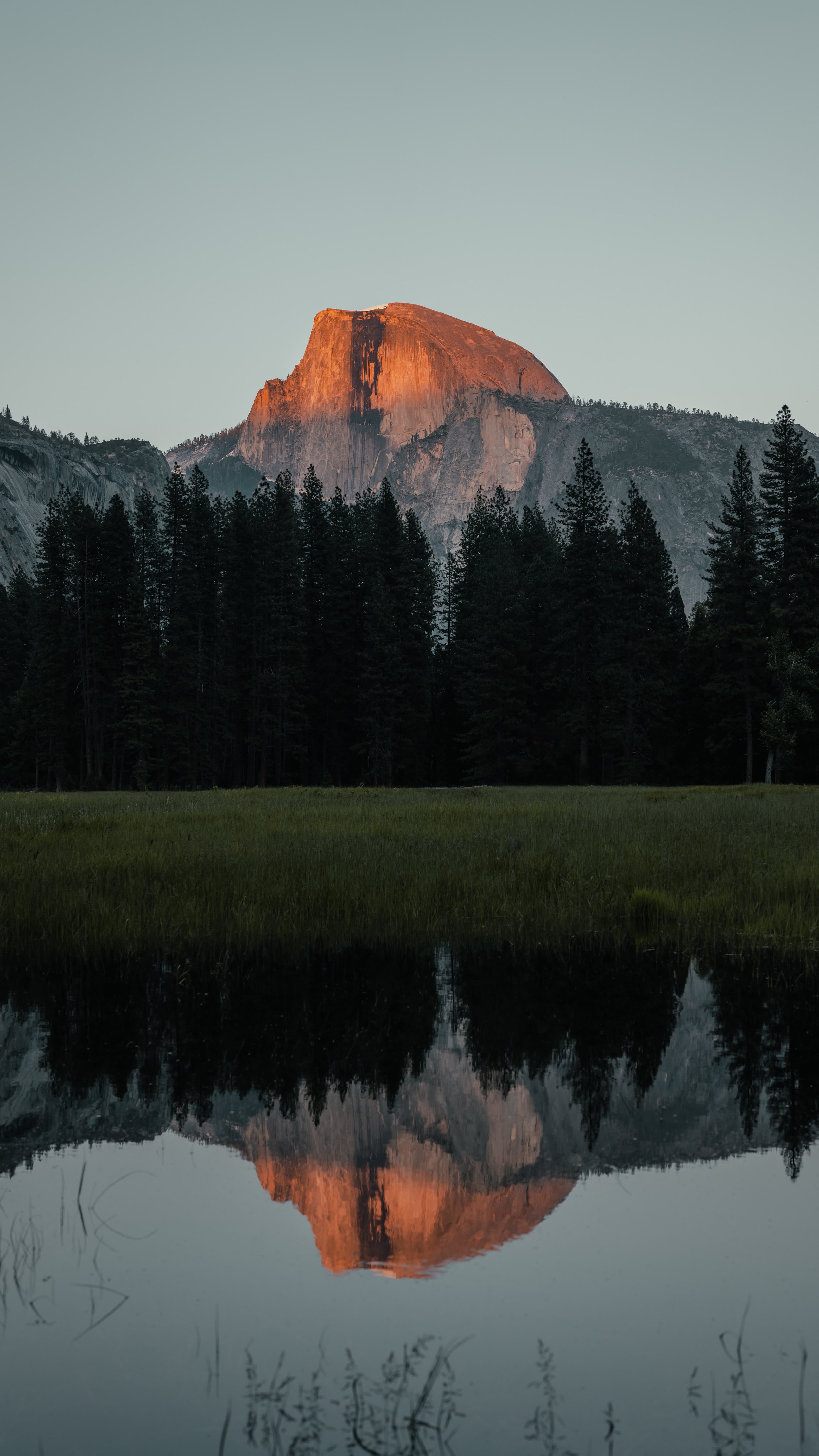 Sunrise or sunset illuminating the top of a large granite mountain, reflected in a calm body of water with green grass and trees in the foreground.