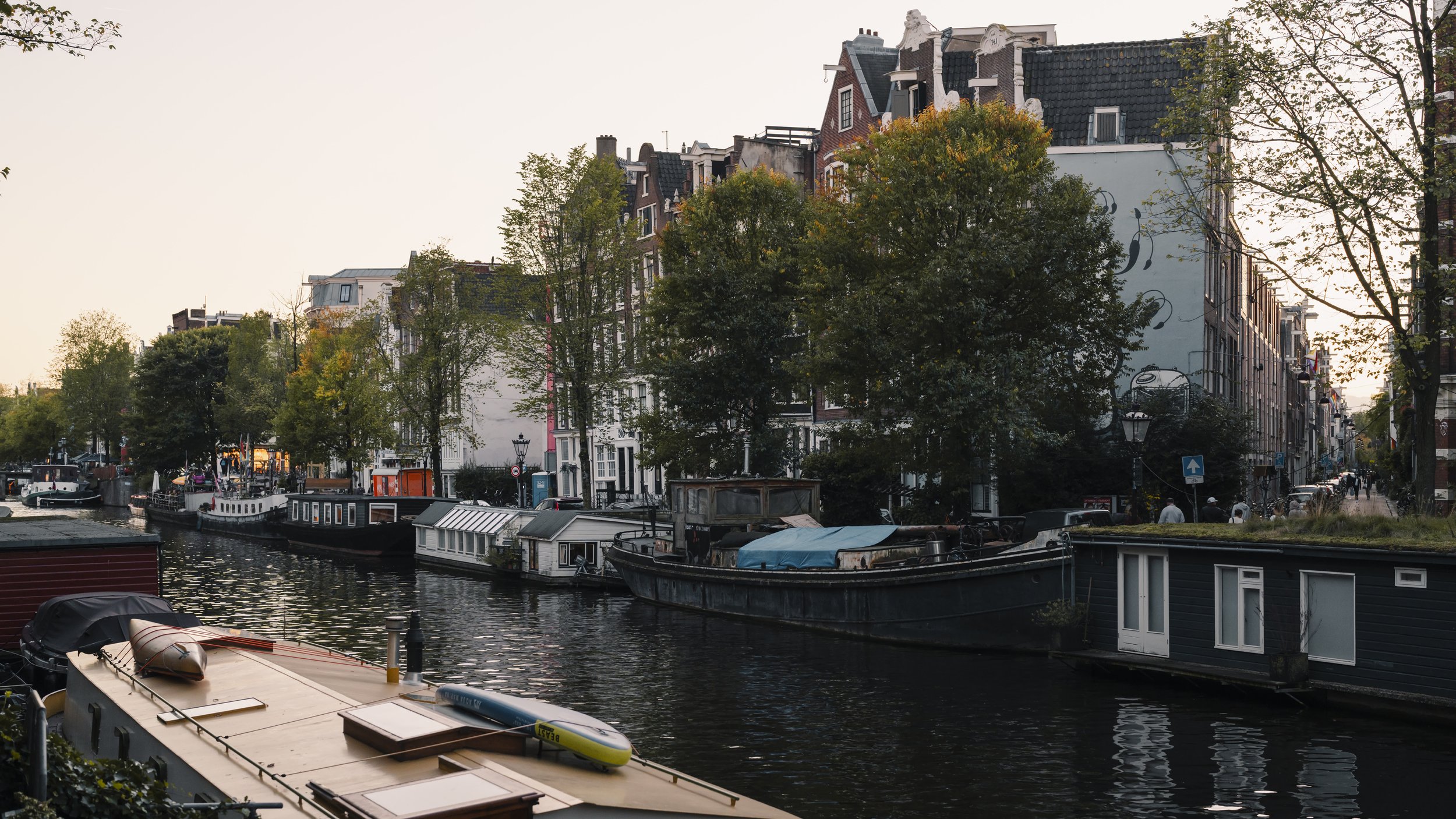 A canal with several boats and houseboats moored along the water, lined with tall trees and narrow buildings with European architectural style.
