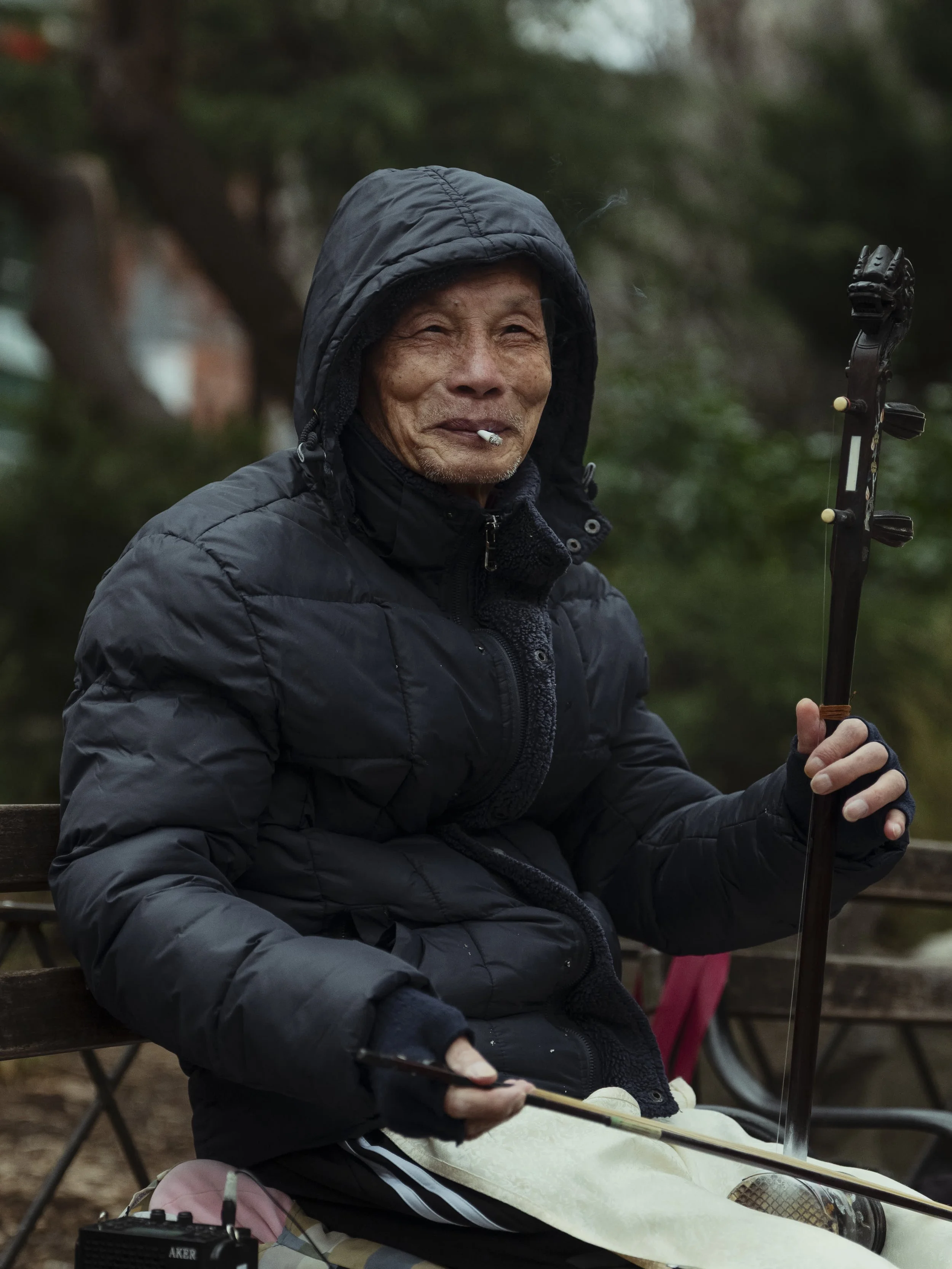 An elderly Asian man sitting outdoors on a park bench, dressed in a black hooded jacket, holding a musical instrument, with a cigarette in his mouth, and surrounded by greenery.
