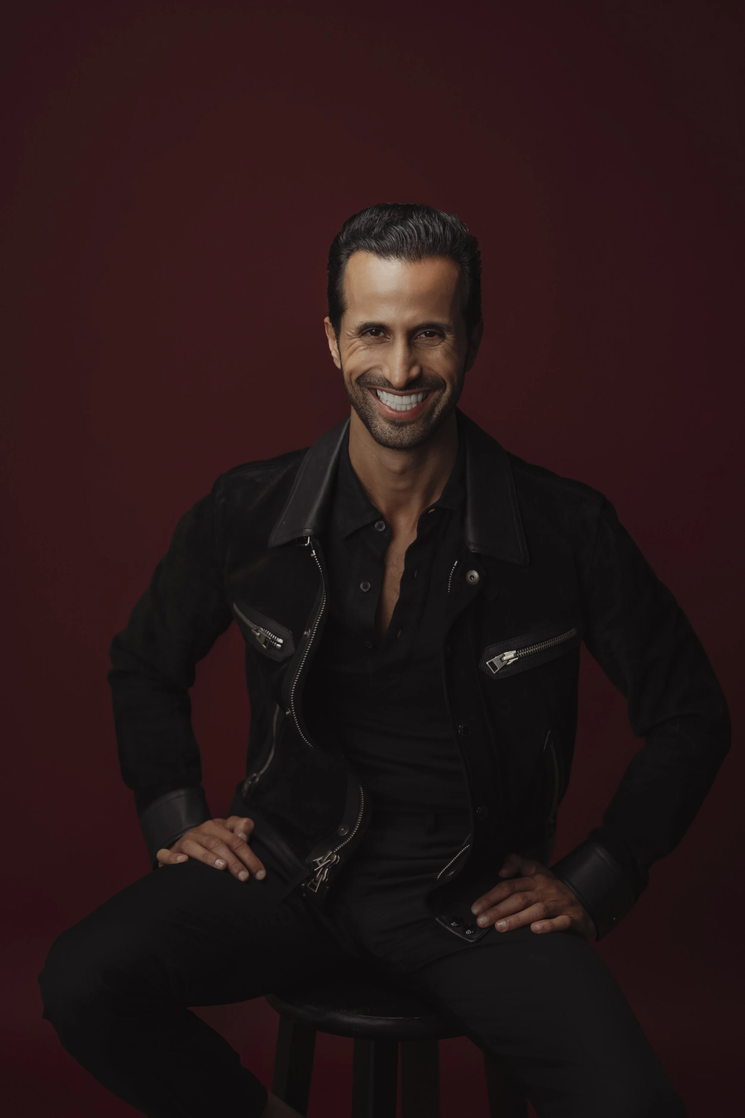 A smiling man with dark hair, dressed in a black leather jacket and black shirt, sitting on a stool against a dark red background.