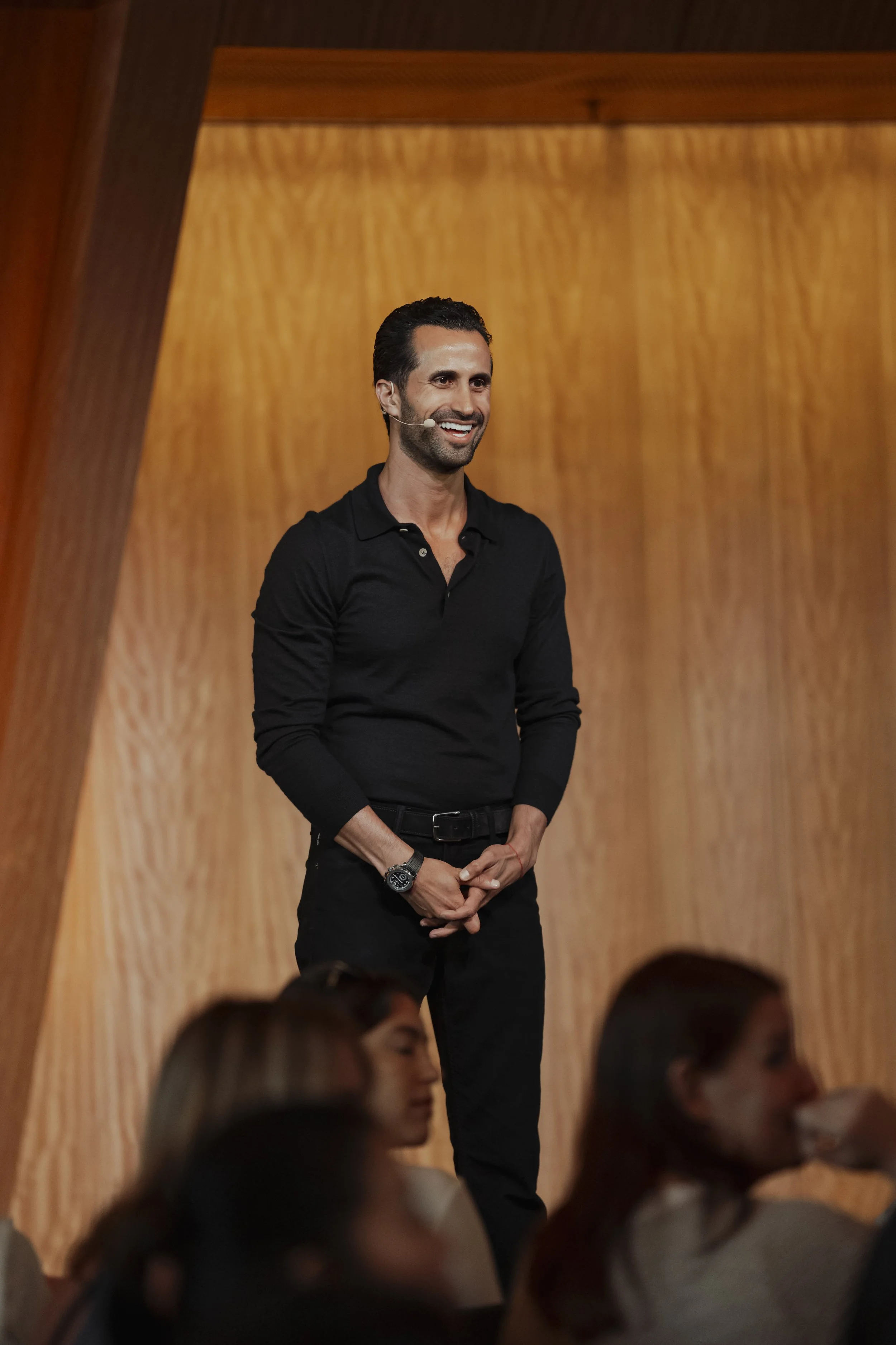 A man in a black long-sleeve polo shirt standing on a stage, smiling, with a wooden background, facing an audience.