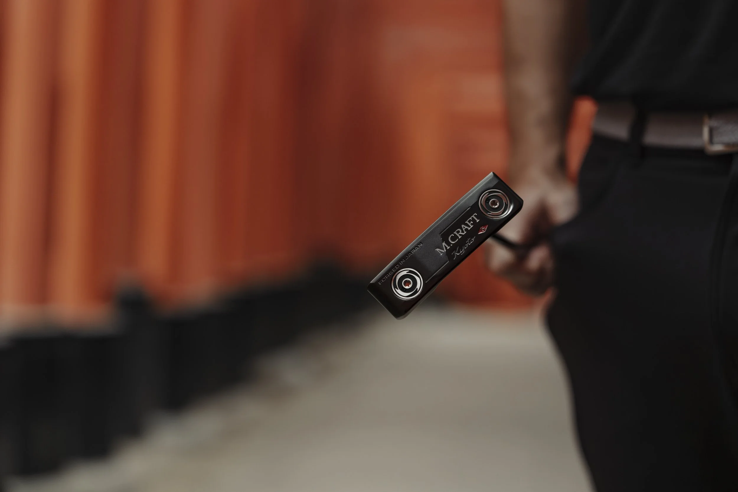 A person in black clothing and glasses holds a black lipstick tube with the label 'M.CRAFT' and 'Kiss' visible, with orange a wooden fence in the background.