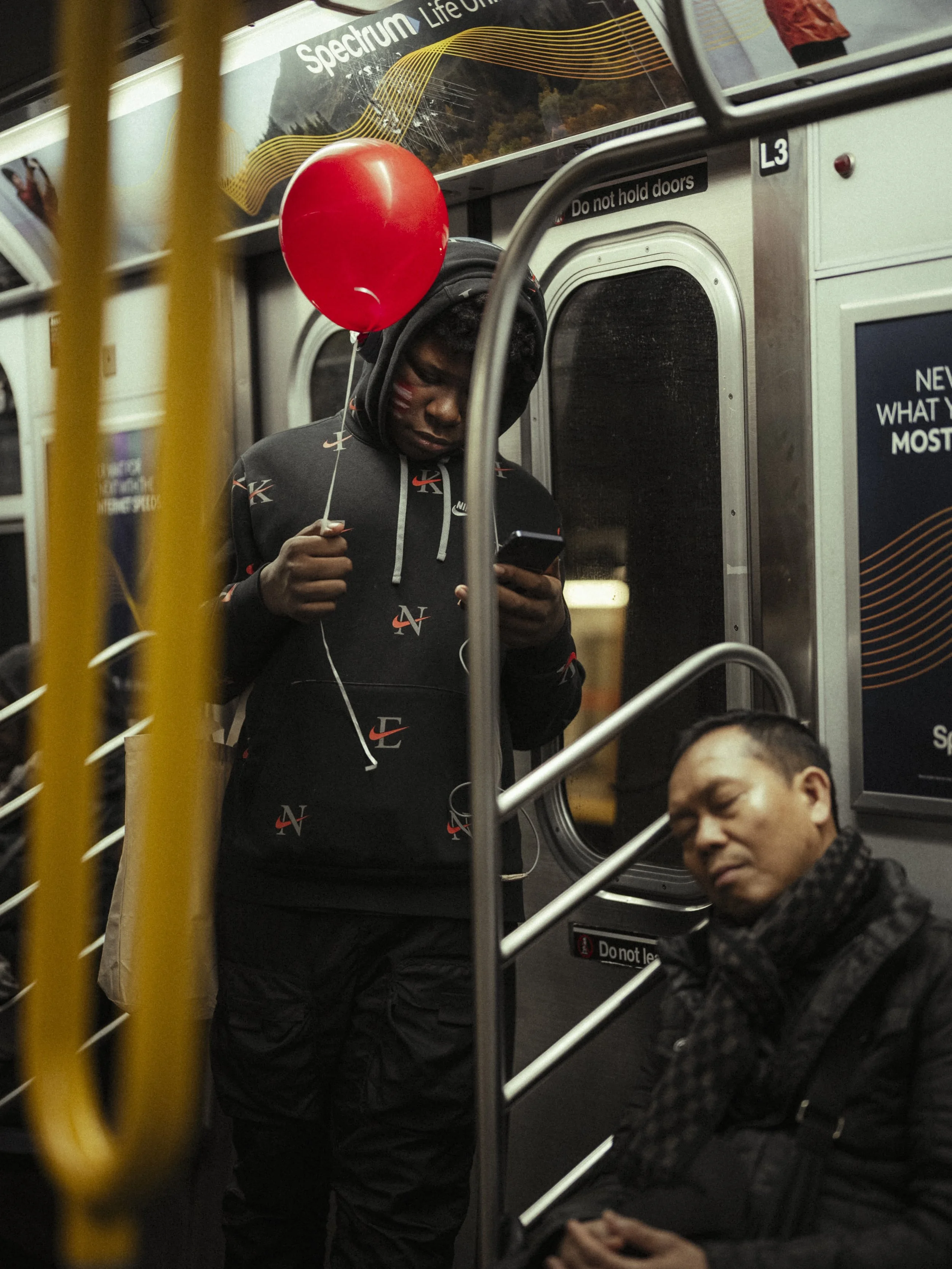 A young man standing inside a subway train, wearing a black hoodie with a pattern and listening to a phone with white earphones, holding a red balloon. Another man with a black scarf and jacket is seated, sleeping or resting with eyes closed.