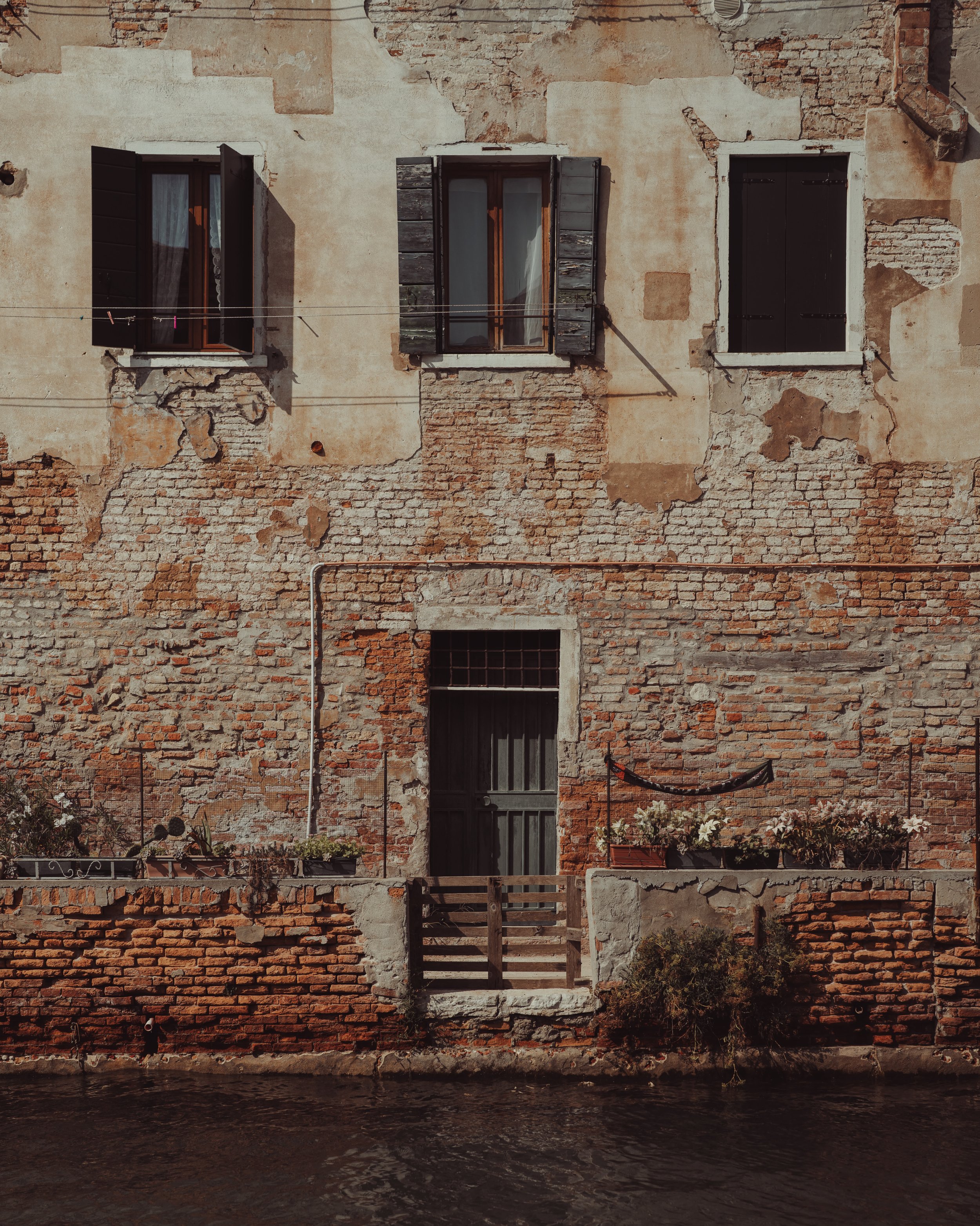Old brick building with three windows, some with open shutters, and a wooden door near the water.