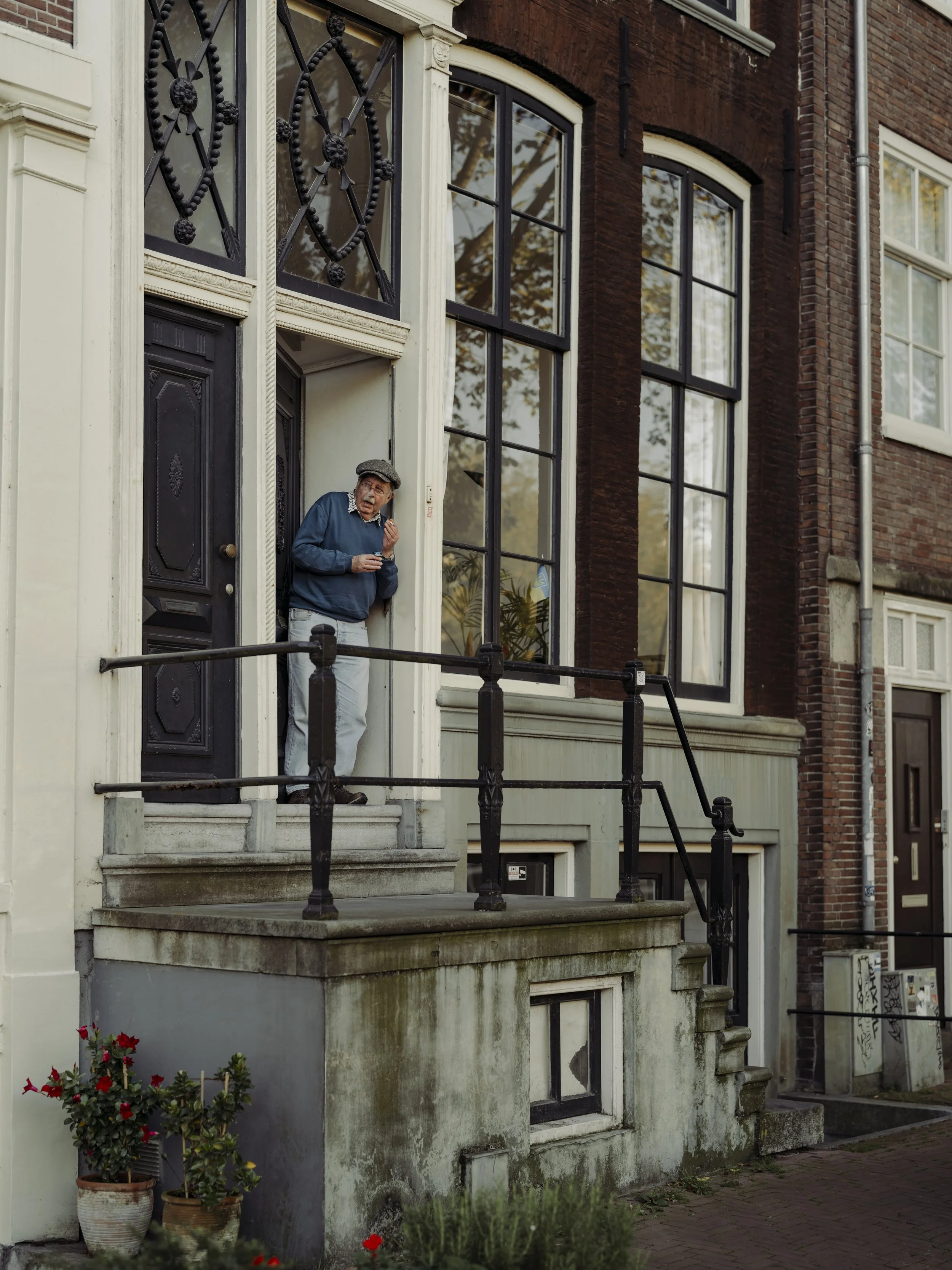 A man leaning outside a house doorway, holding a cup, wearing a blue sweater, light-colored pants, and a cap, standing on steps in front of a large window with black framing.