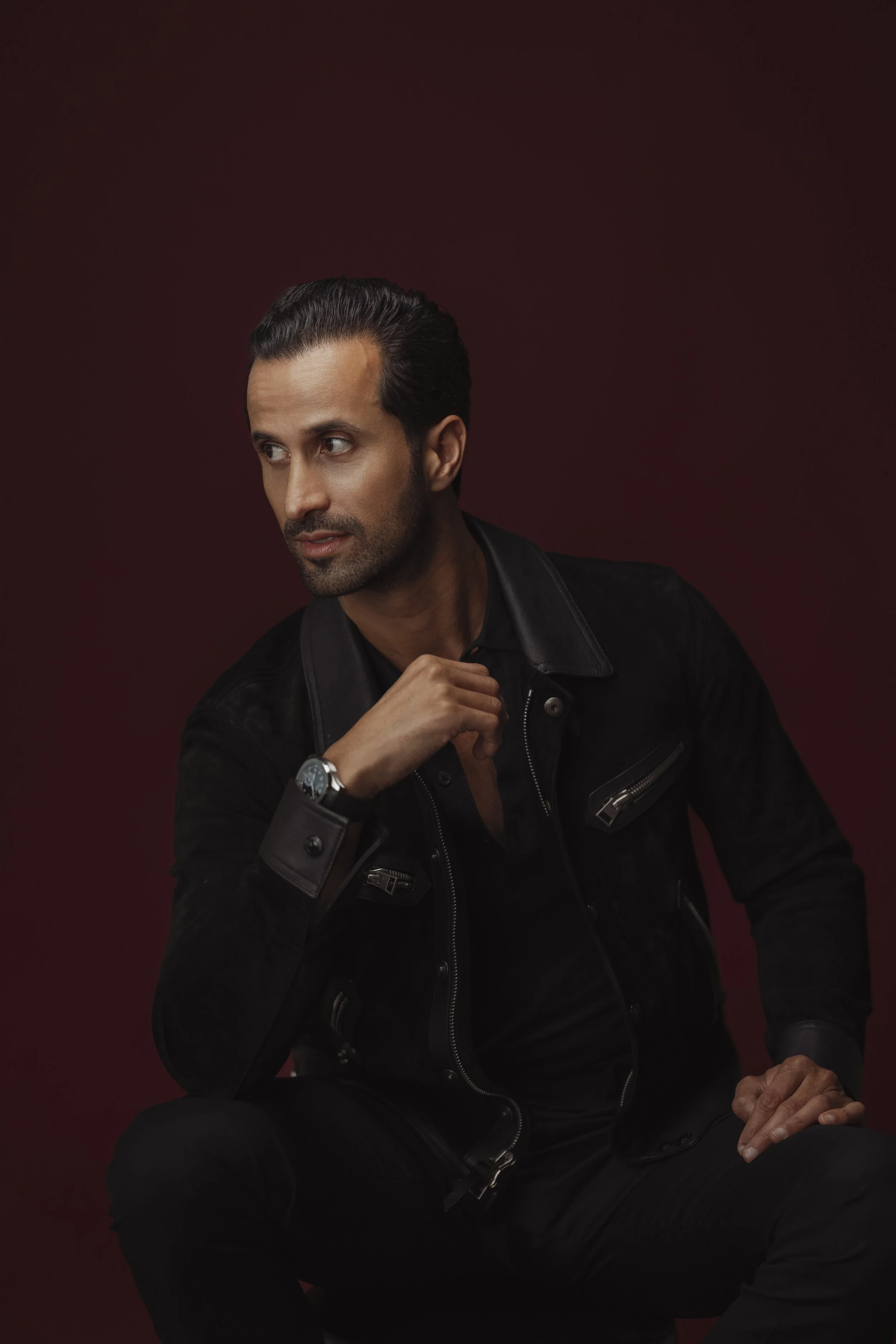 A man with dark hair and a beard wearing a black leather jacket, sitting against a dark red background, looking to the side with a thoughtful expression.