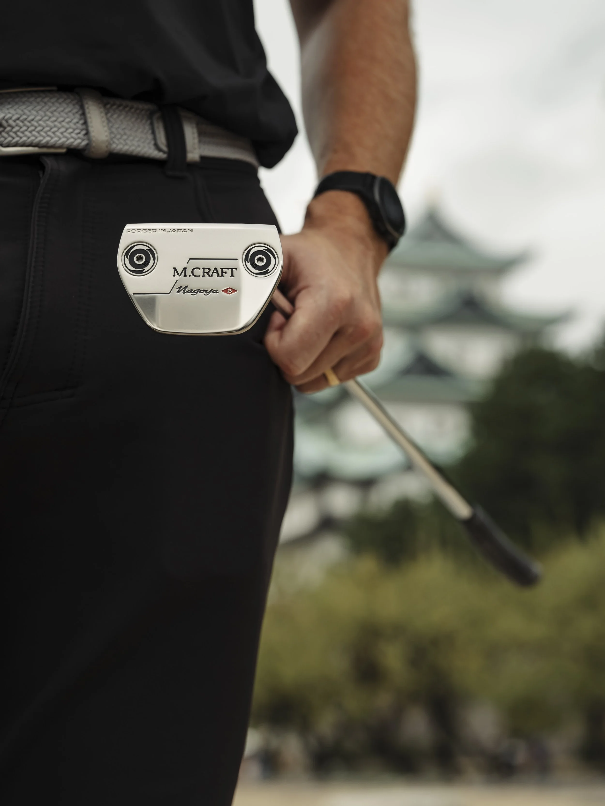 Person holding a golf club with a white golf club head visible, wearing black pants, a gray belt, and a black watch, with a background of a pagoda-style building and trees.