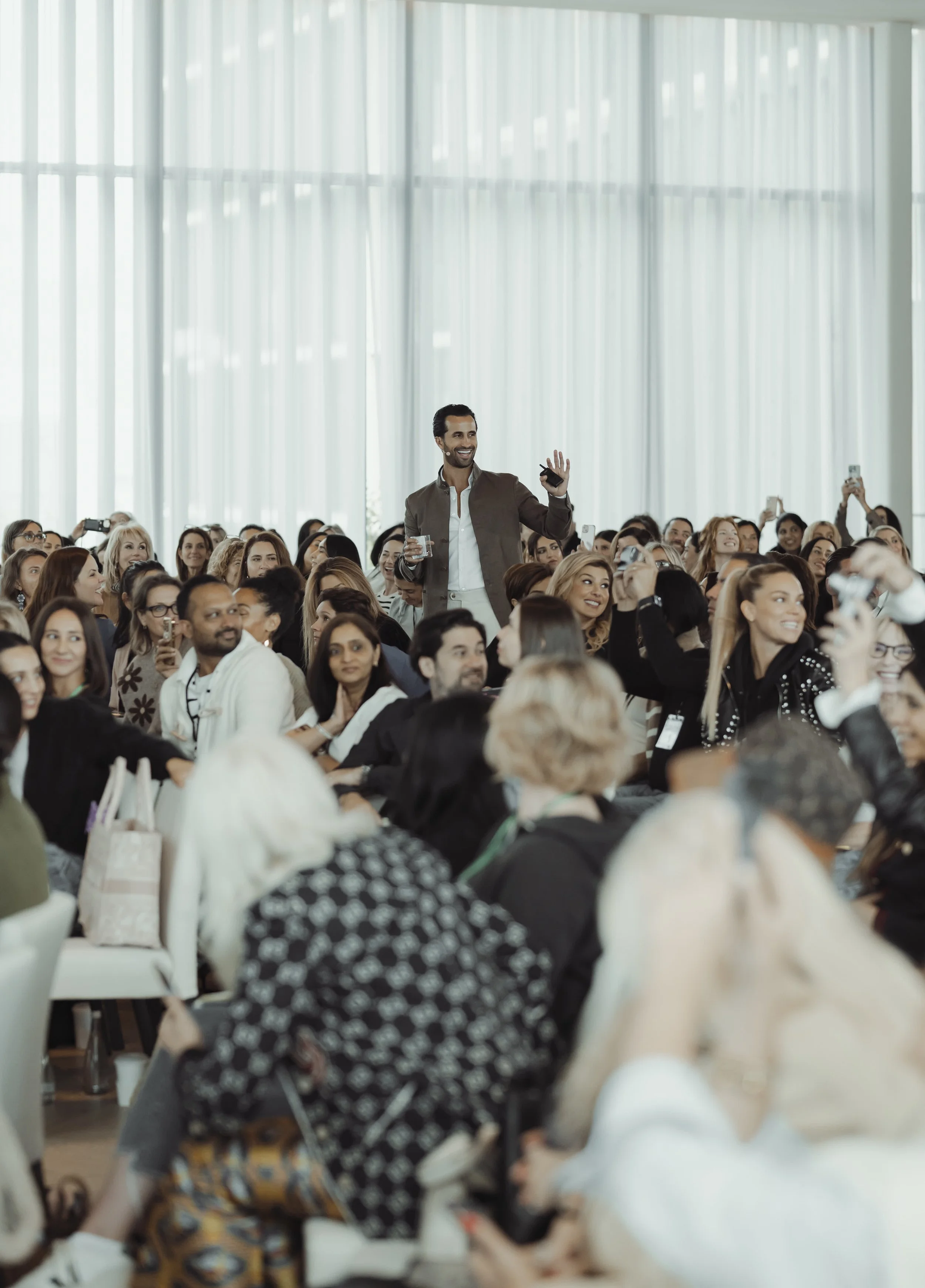 A large group of people seated indoors during a conference or seminar, with a man standing and smiling while waving.