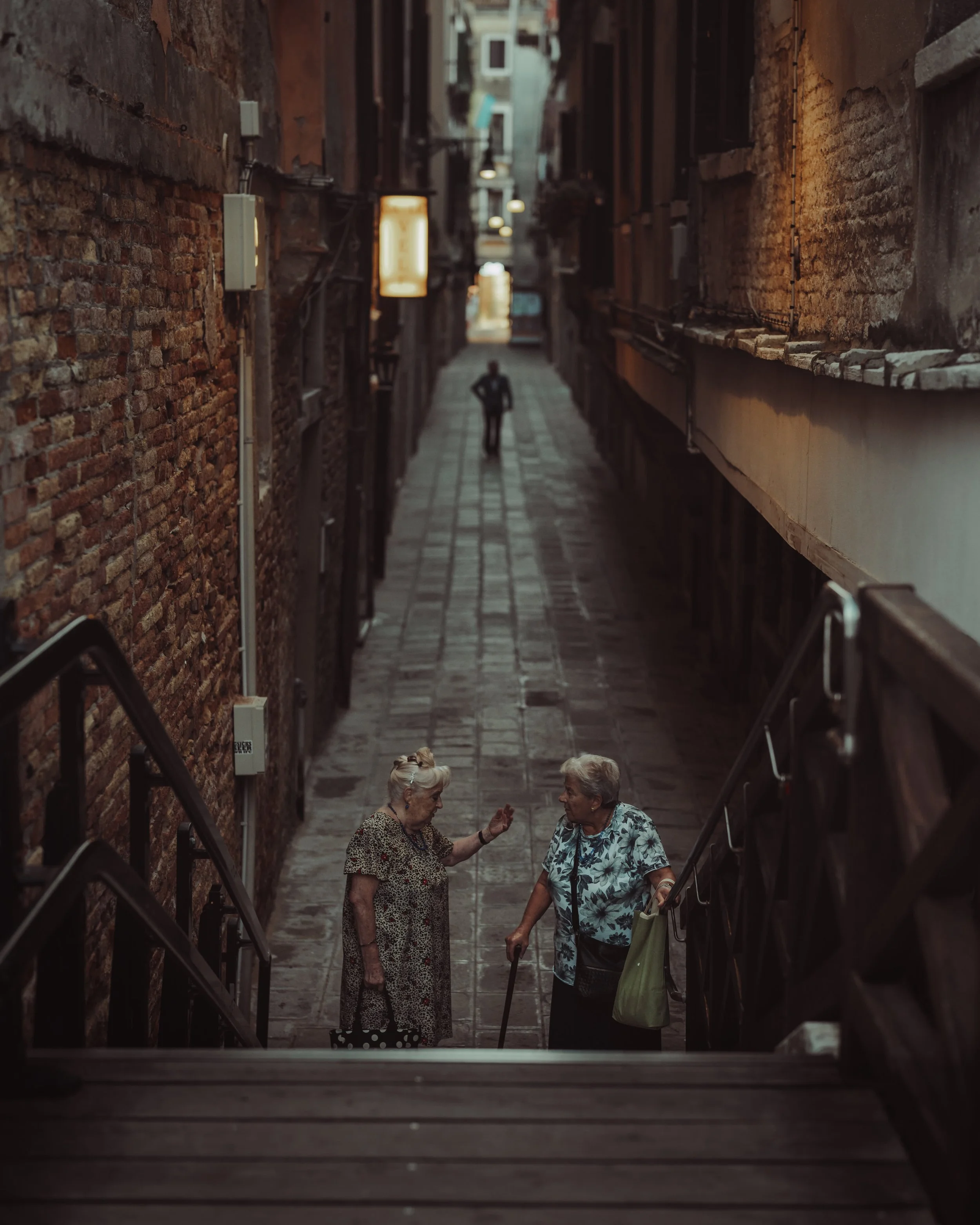 Two elderly women are talking on the stairs in a narrow alley with brick walls on either side.
