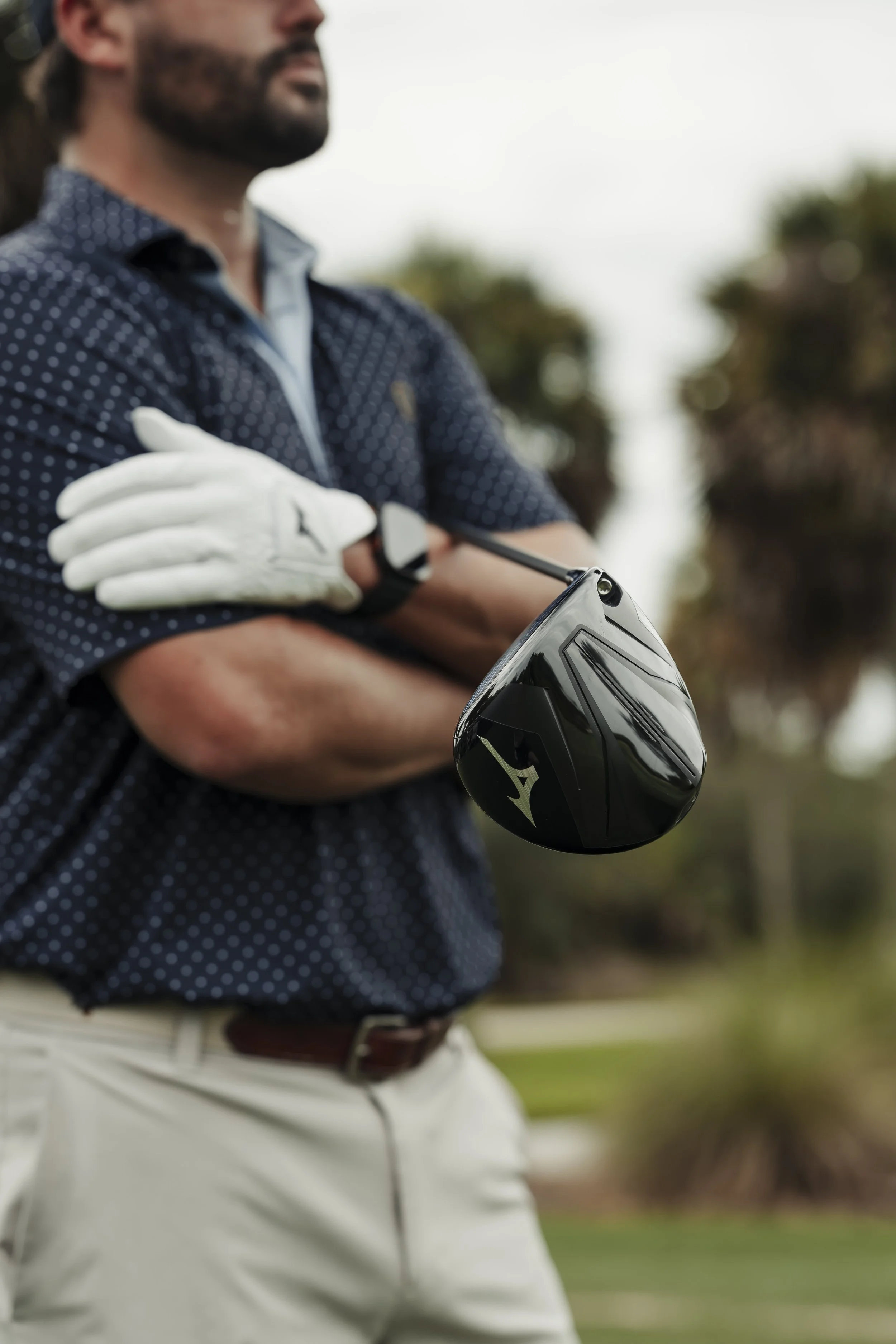 A man standing outdoors holding a golf club with crossed arms, wearing a navy polo shirt, khaki pants, a belt, white golf glove on one hand, and smartwatch.