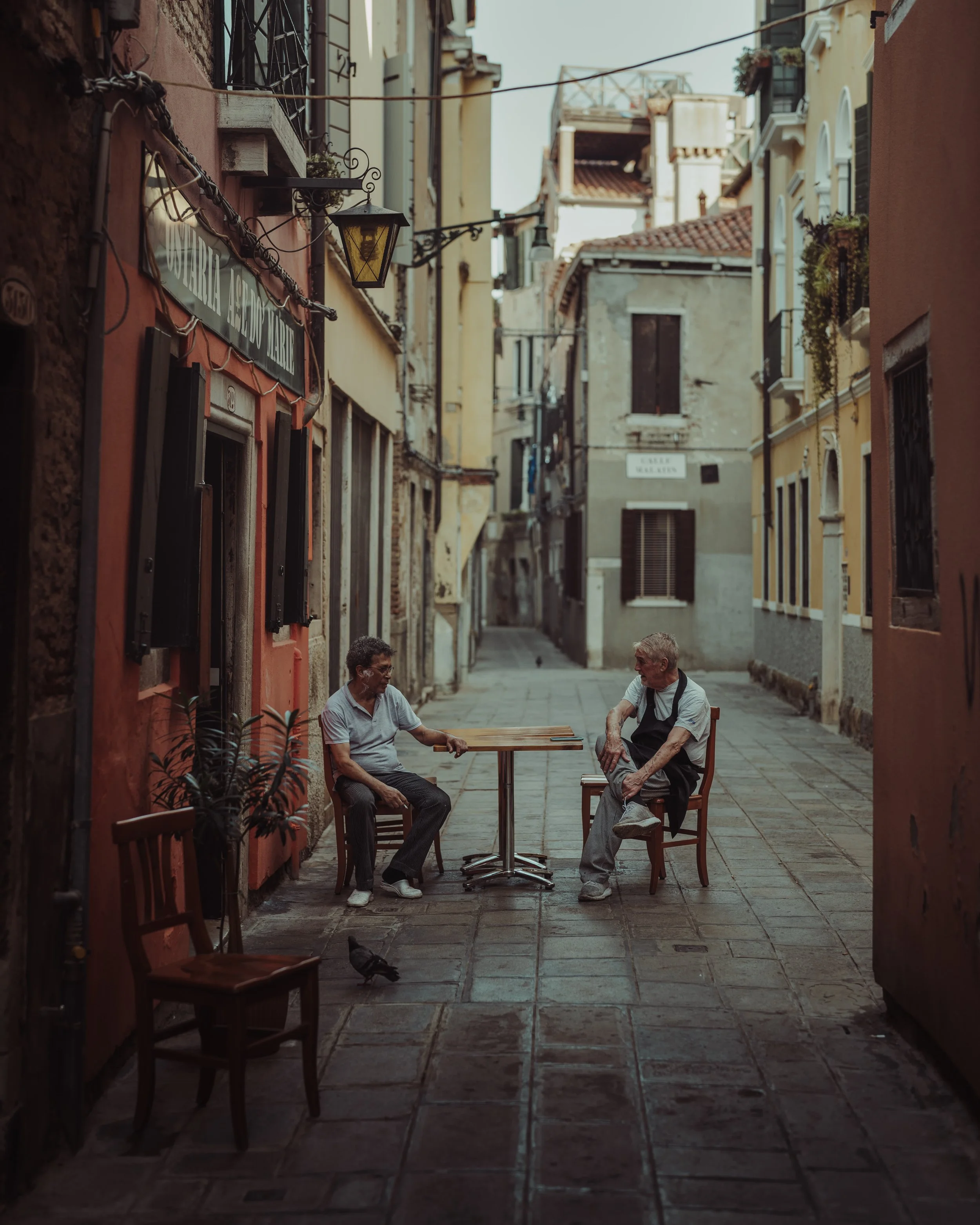 Two elderly men sitting and talking at an outdoor table on a narrow European street with colorful buildings and a pigeon on the ground.