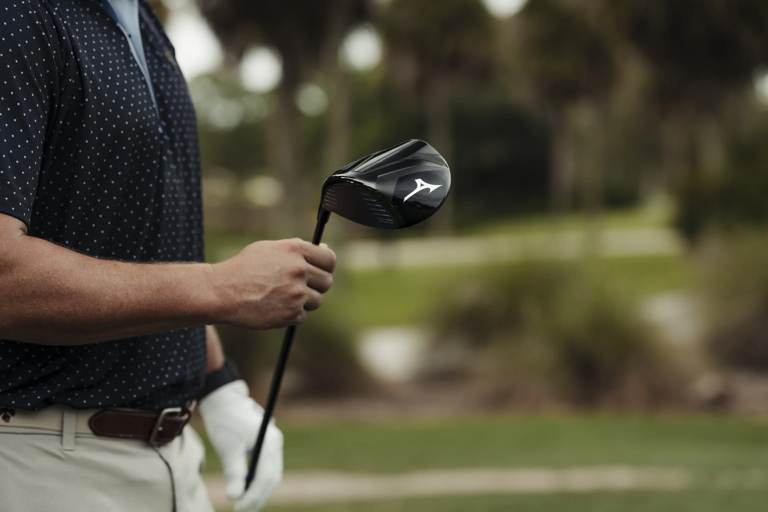 A person on a golf course holding a black golf club driver with a white logo, wearing a black polka dot shirt and beige pants.