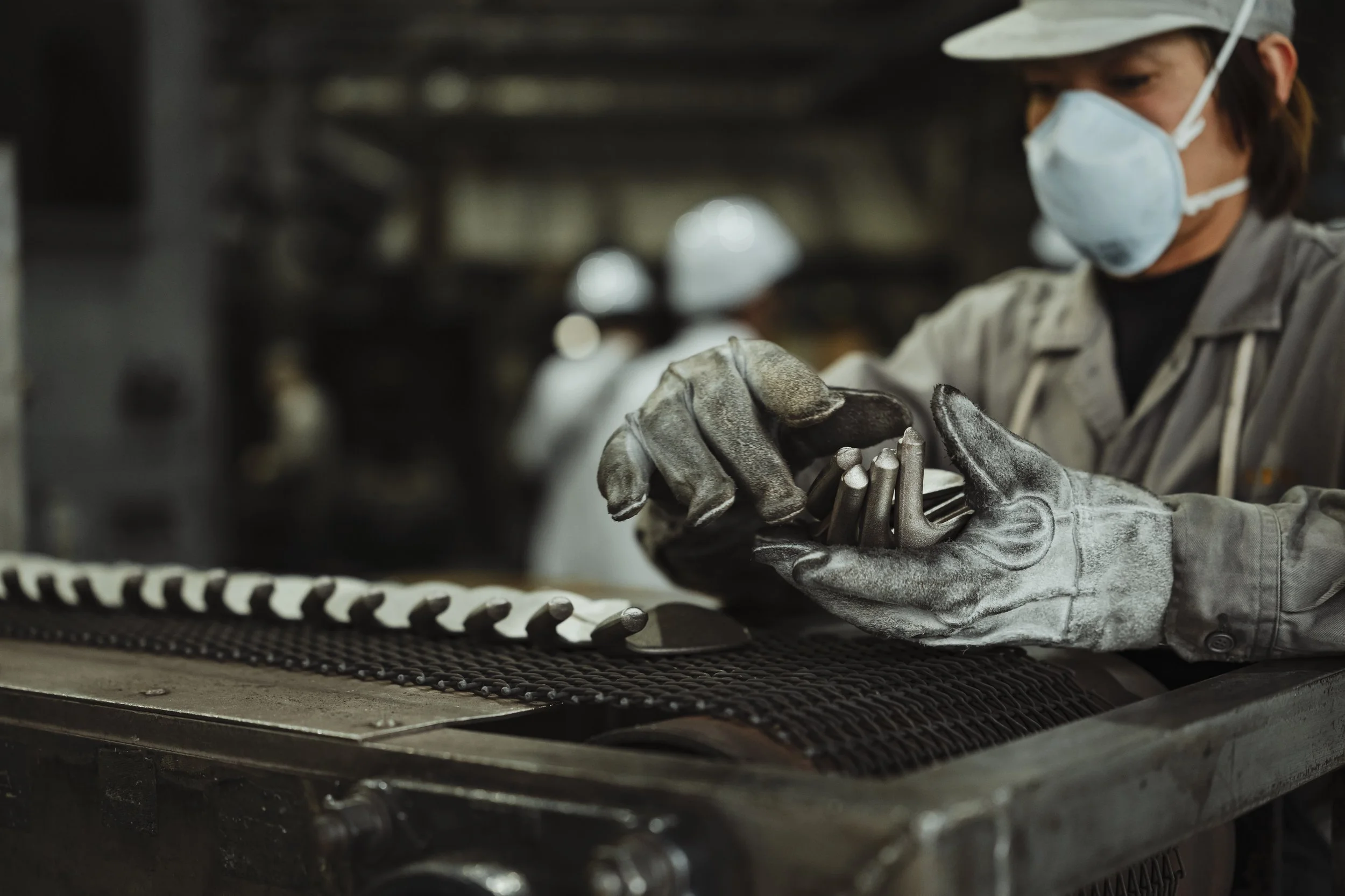 A worker in a factory wearing gloves, a face mask, and a hat, handling a metal component on a workbench.