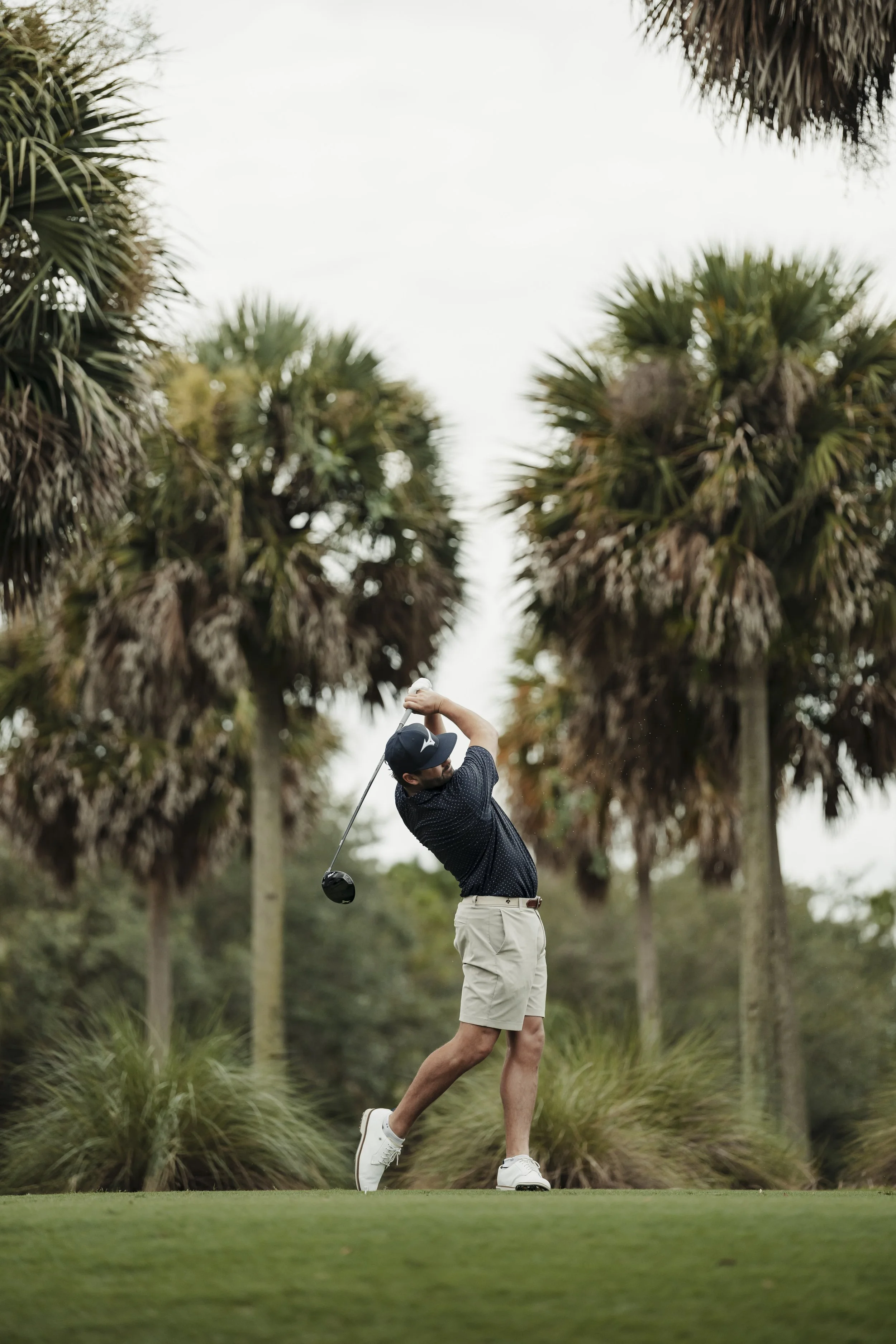 A man playing golf on a golf course, swinging a club amidst tall palm trees with a cloudy sky overhead.