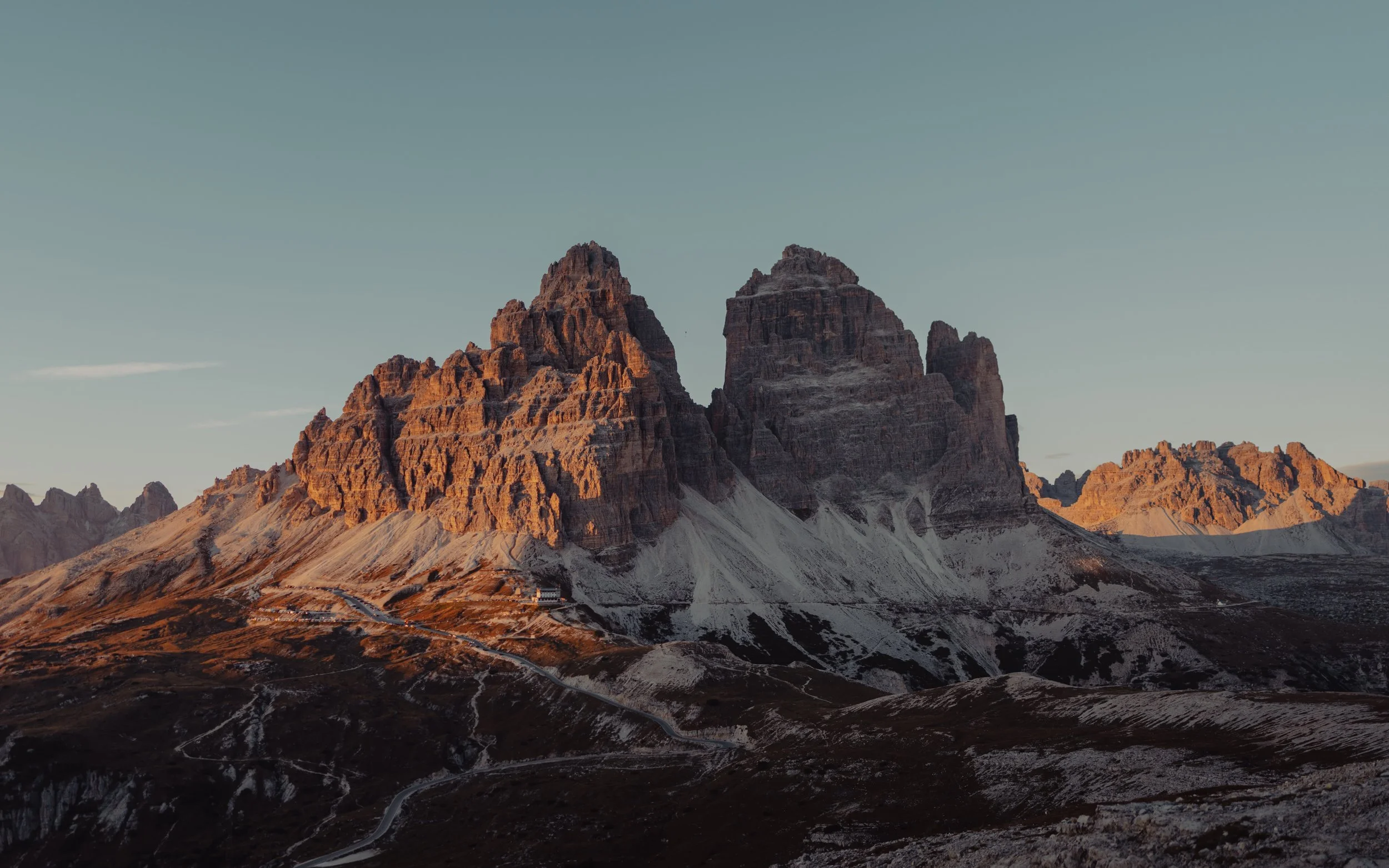 Mountain peaks at sunset with snow patches and winding roads on the landscape.