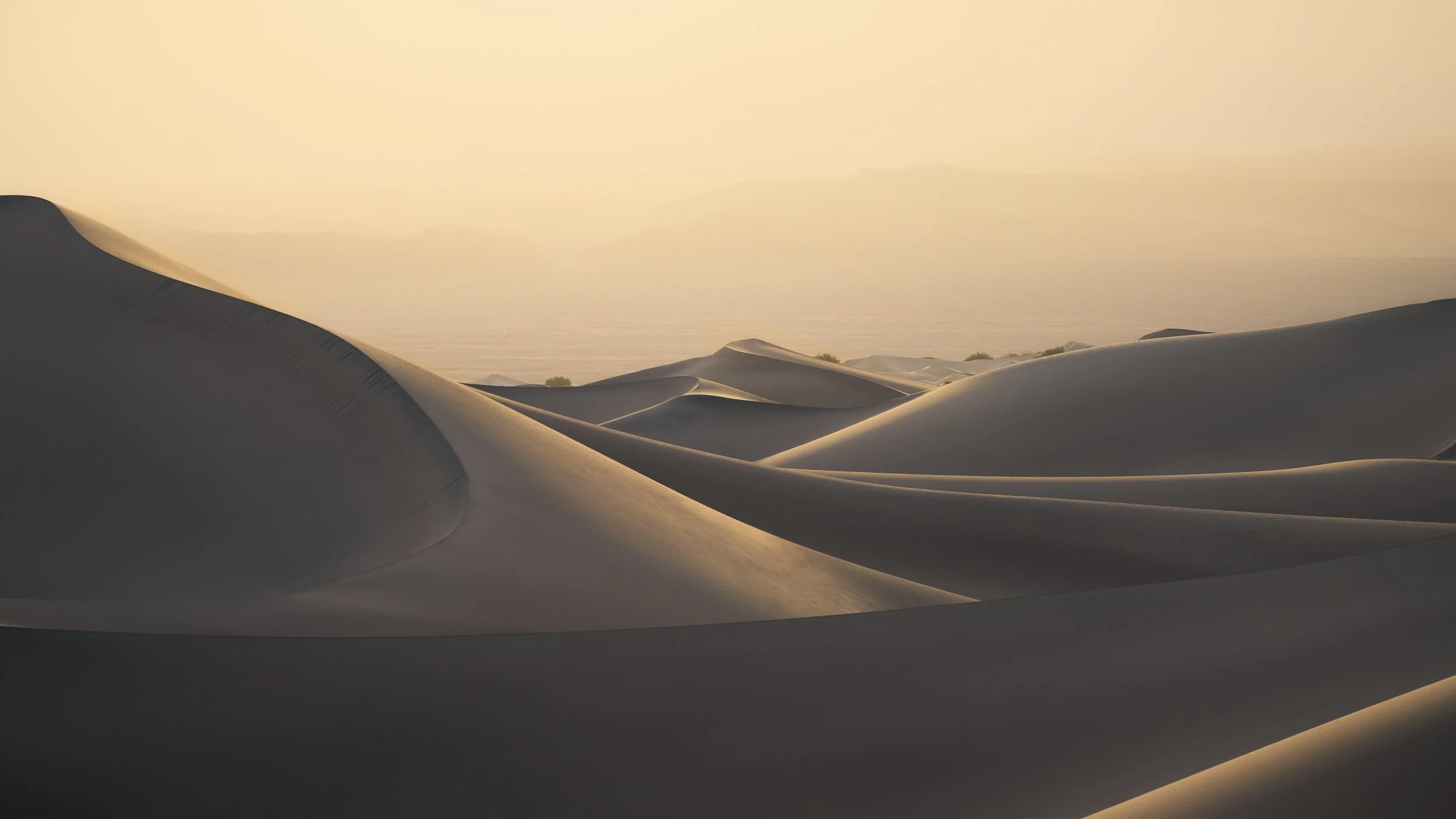 Sand dunes in a desert during sunset with a hazy sky and distant mountains.