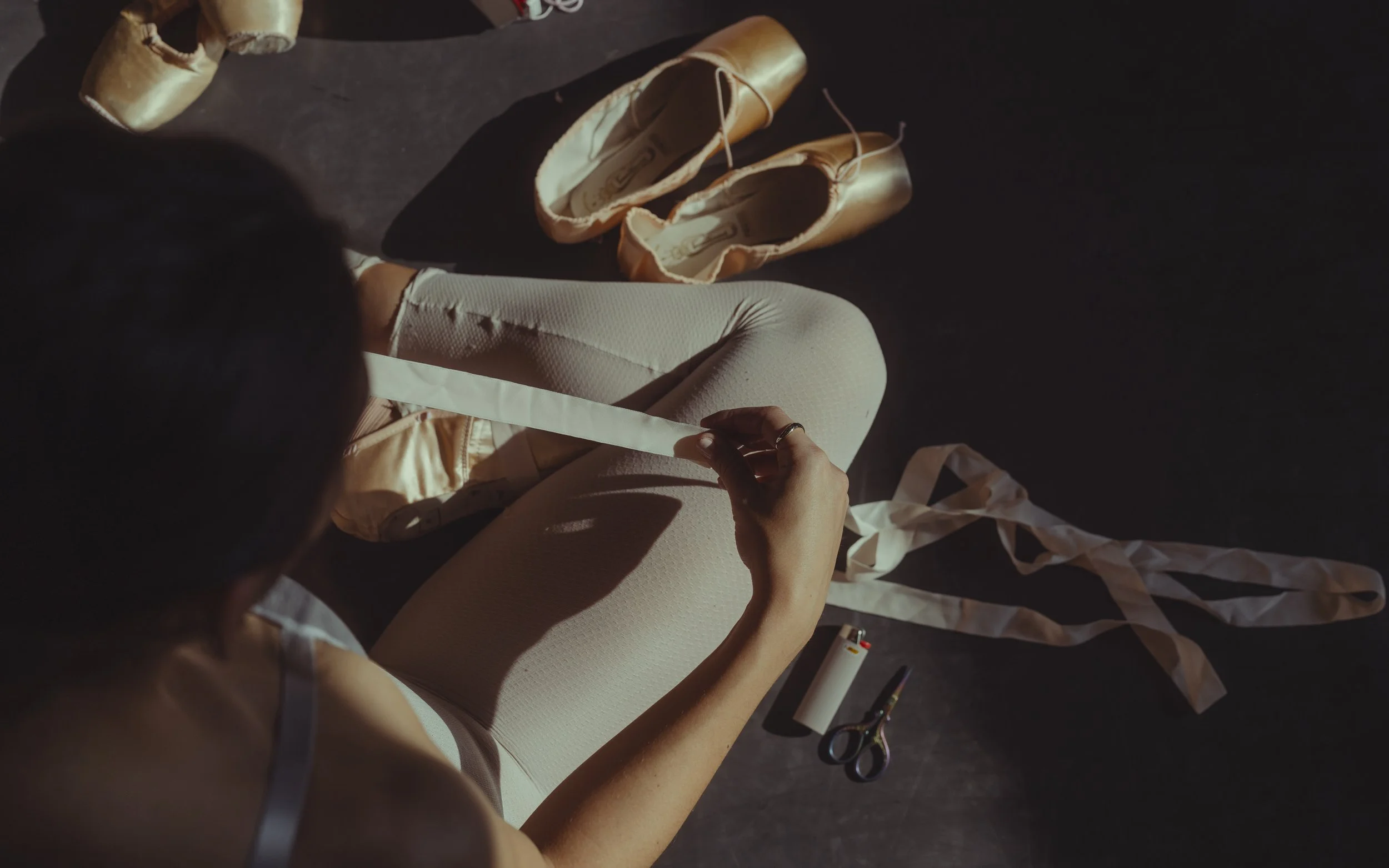 A ballet dancer sitting on the floor with ballet shoes and tutu, preparing for a dance, with scissors, a lighter, and a roll of white ribbon nearby.