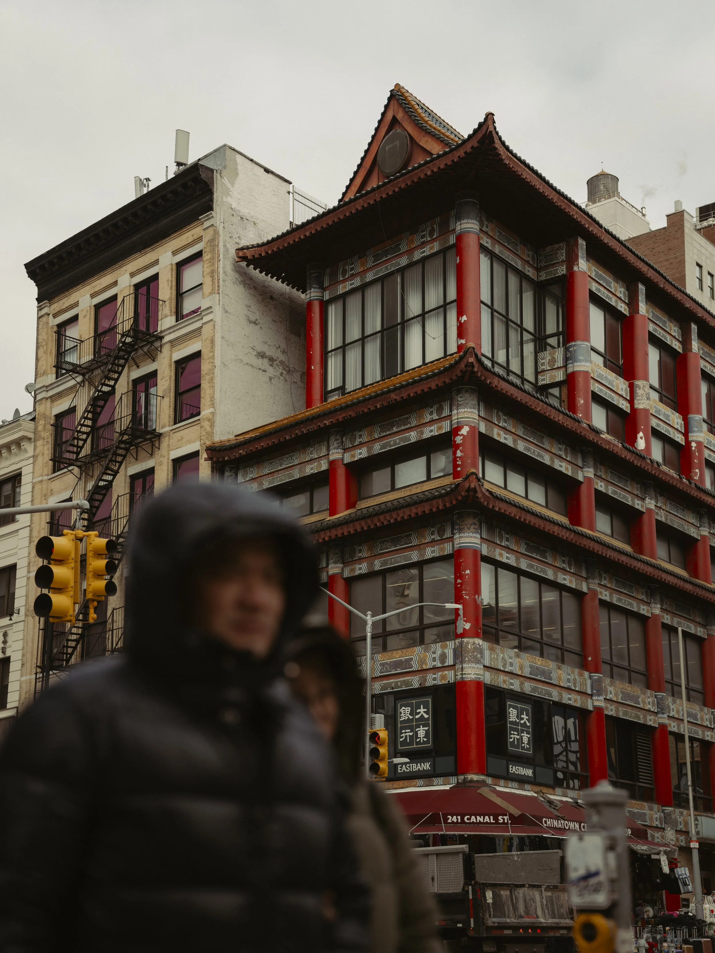 A traditional Chinese-style building with red columns and ornate detailing on a city street, with a person in a black jacket with a hood in the foreground.