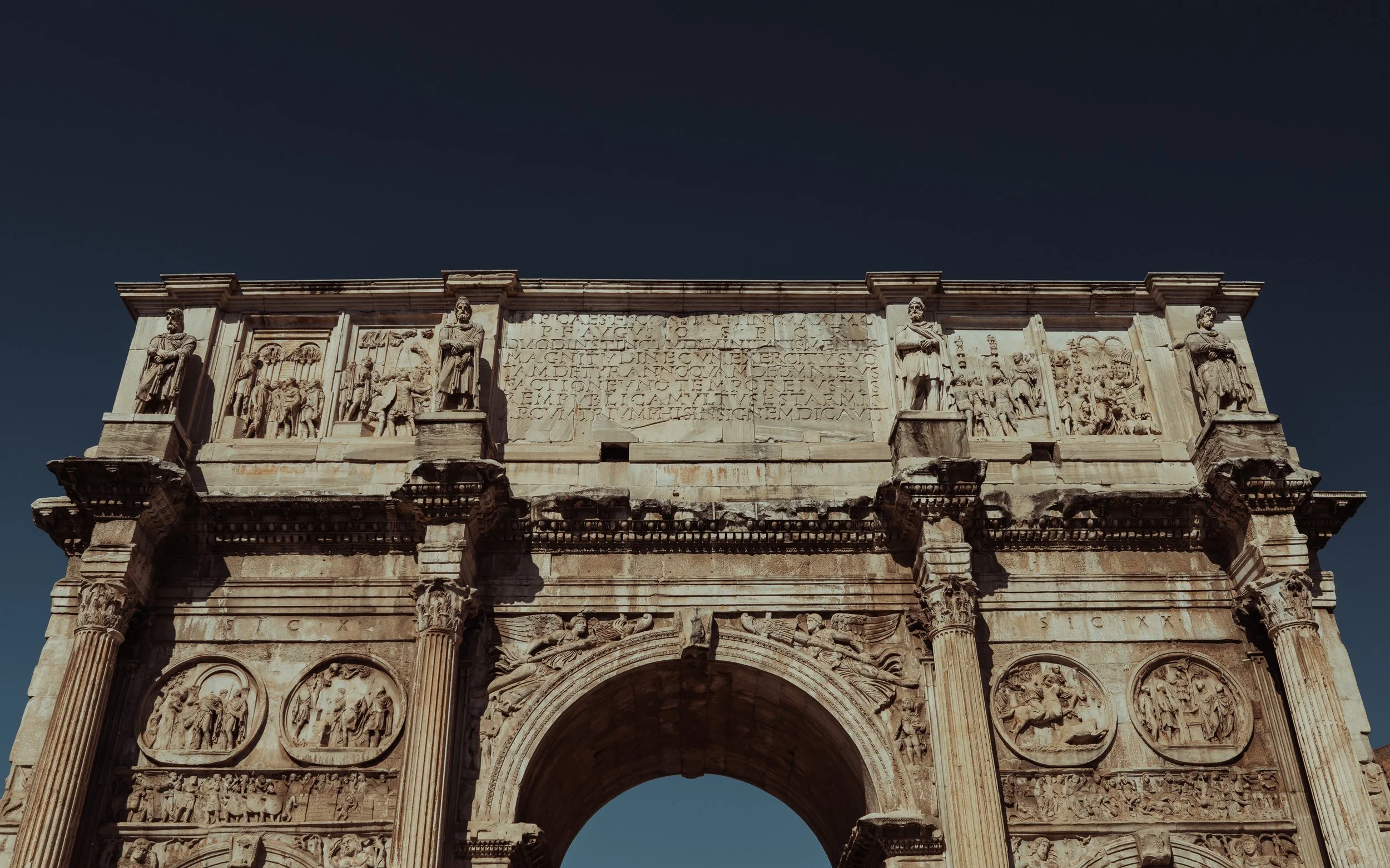 Ancient Roman triumphal arch with detailed carvings, sculptures, and inscriptions, set against a dark sky.
