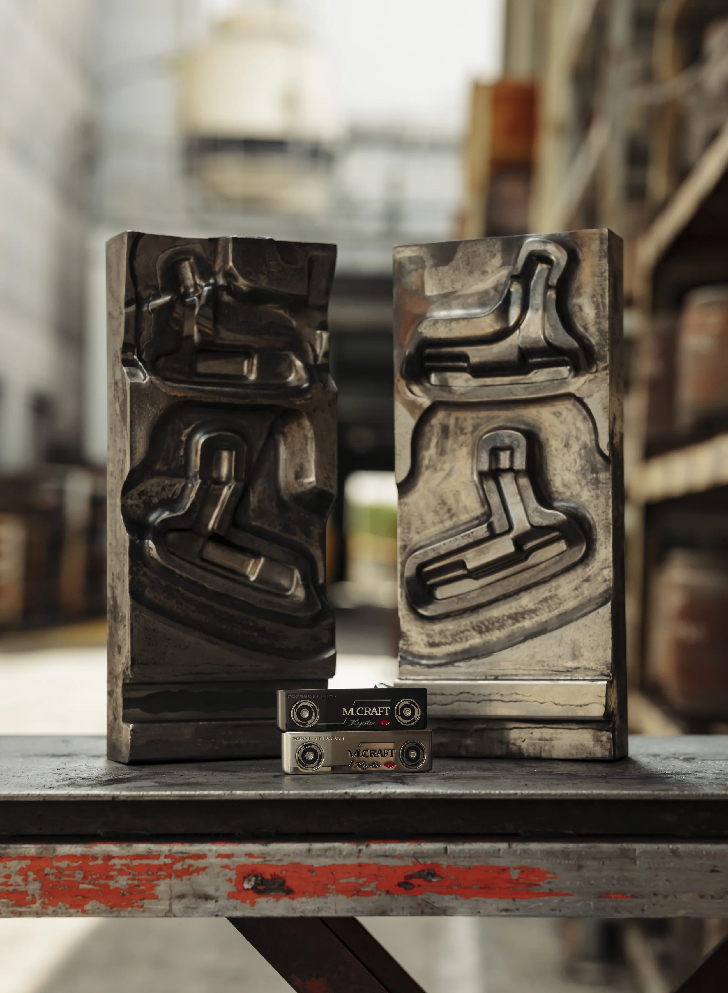 Two metal molds with internal channels for casting, placed on a wooden workbench with two metal objects labeled 'M.CRAFT' in front of them, in a workshop setting.