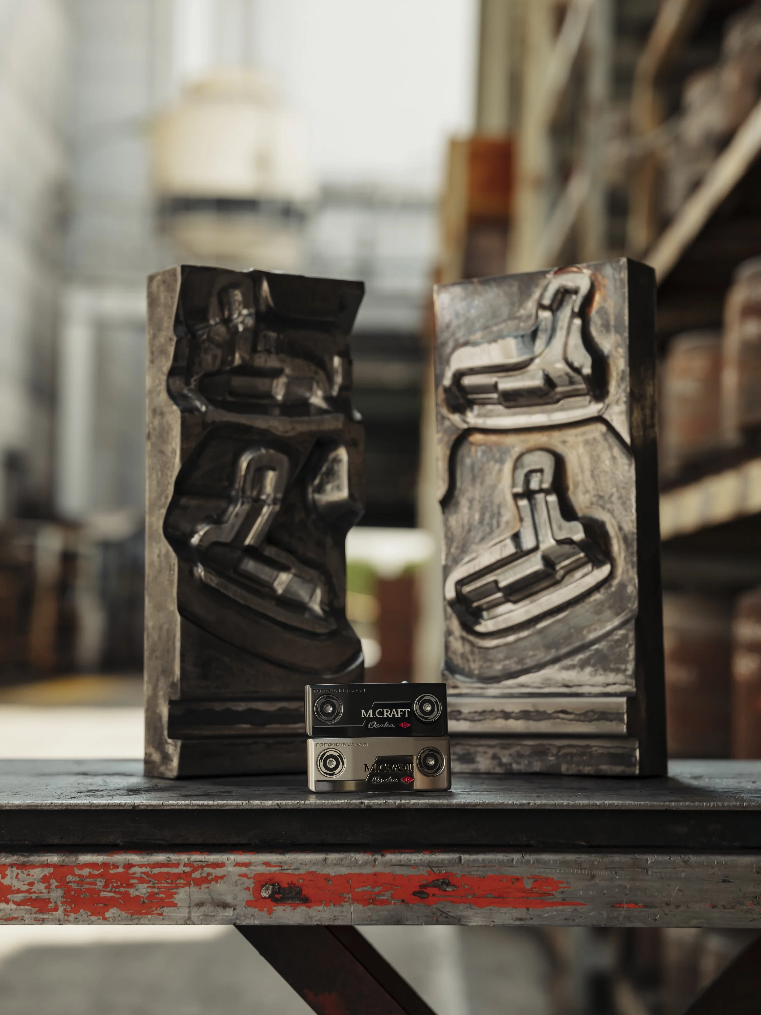 Two metal molds with embedded skateboarding shoes, placed on a wooden table with two small metallic objects labeled 'M. CRAFT Osaka' in front. Background shows shelves with various items in an industrial setting.