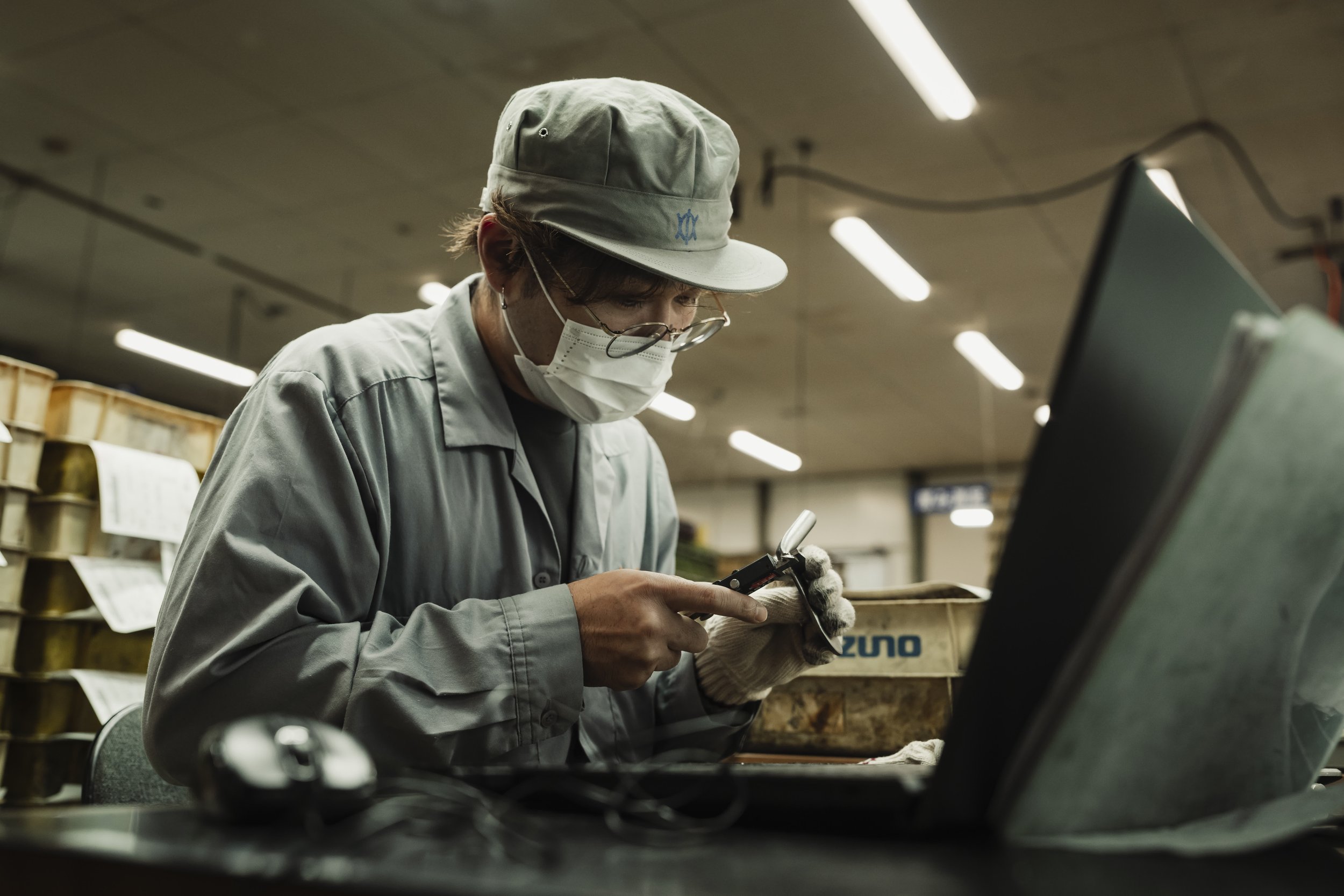 A worker wearing a gray cap, glasses, and a face mask, using a handheld device inside an industrial or warehouse setting.