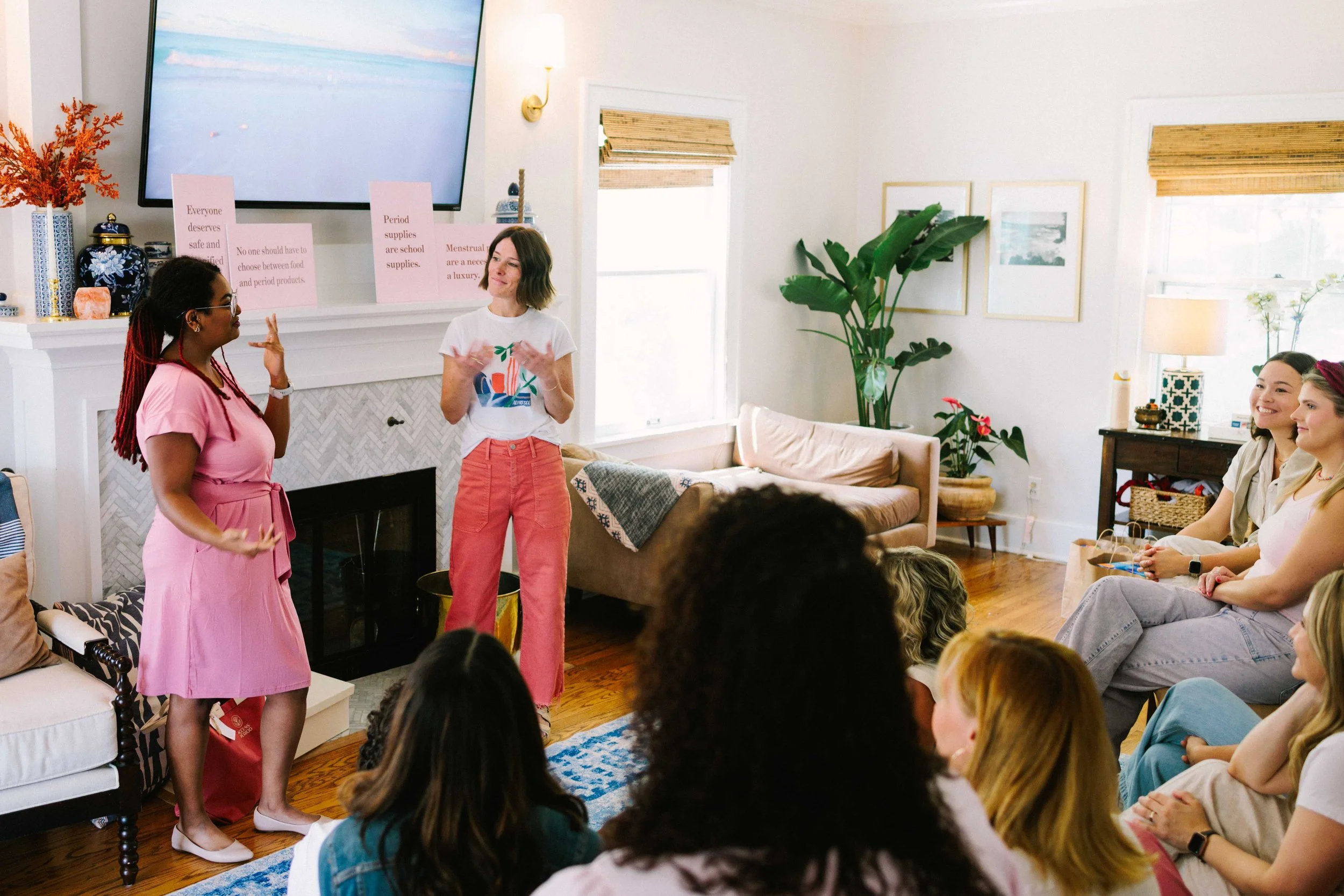 Group of women attending a casual presentation or workshop in a cozy living room, some standing and some sitting, with a large television on the wall and decorative elements including plants and framed pictures.