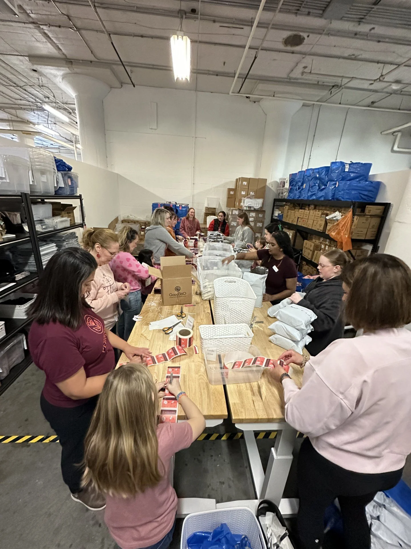 Group of people packing items at a long table in an industrial storage room, with shelves filled with boxes and supplies in the background.