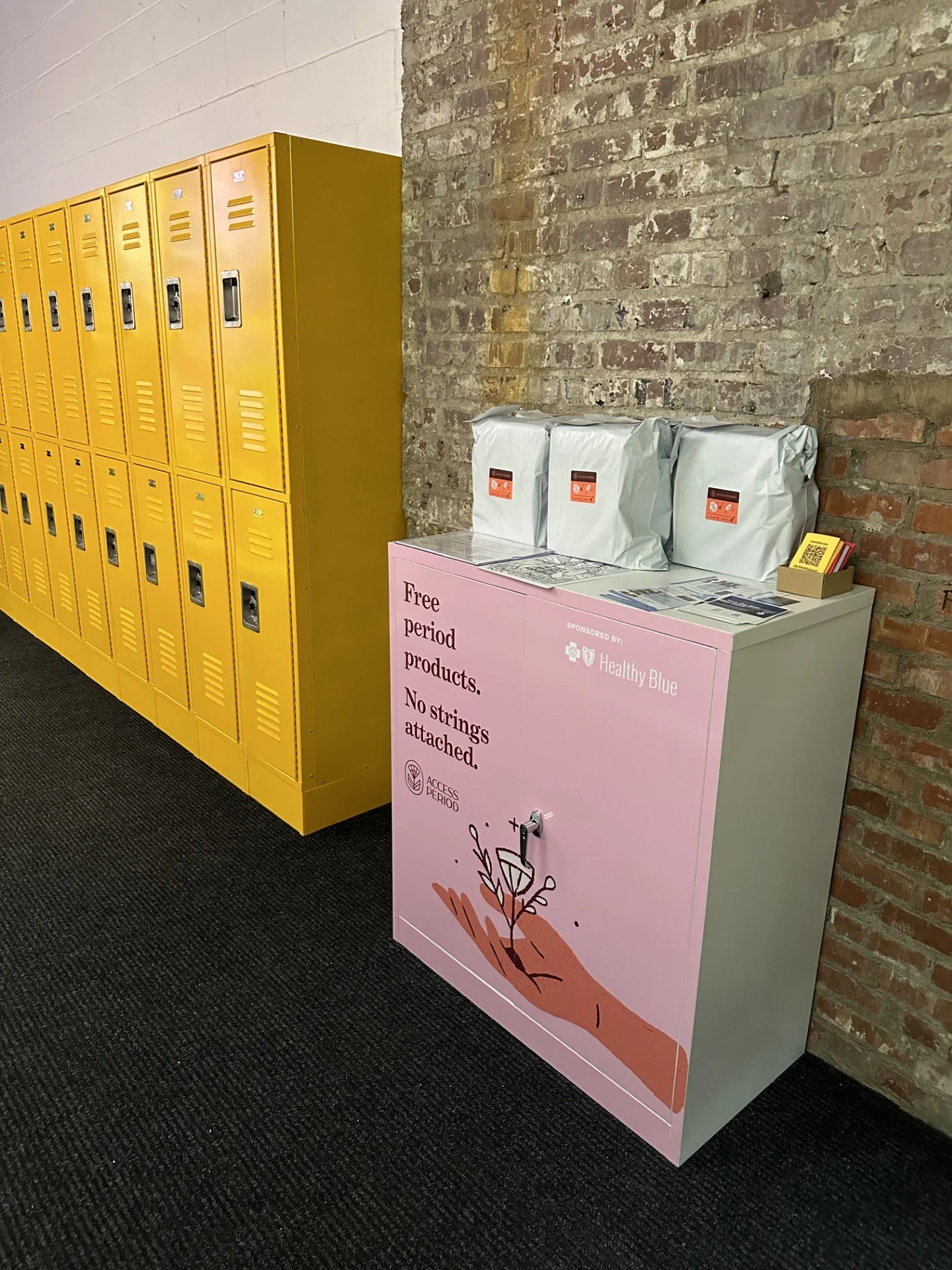 A row of yellow lockers against a white brick wall, next to a pink cabinet that says, "Free Period Products. No Strings Attached." with pamphlets and three white packages on top against a brick wall.