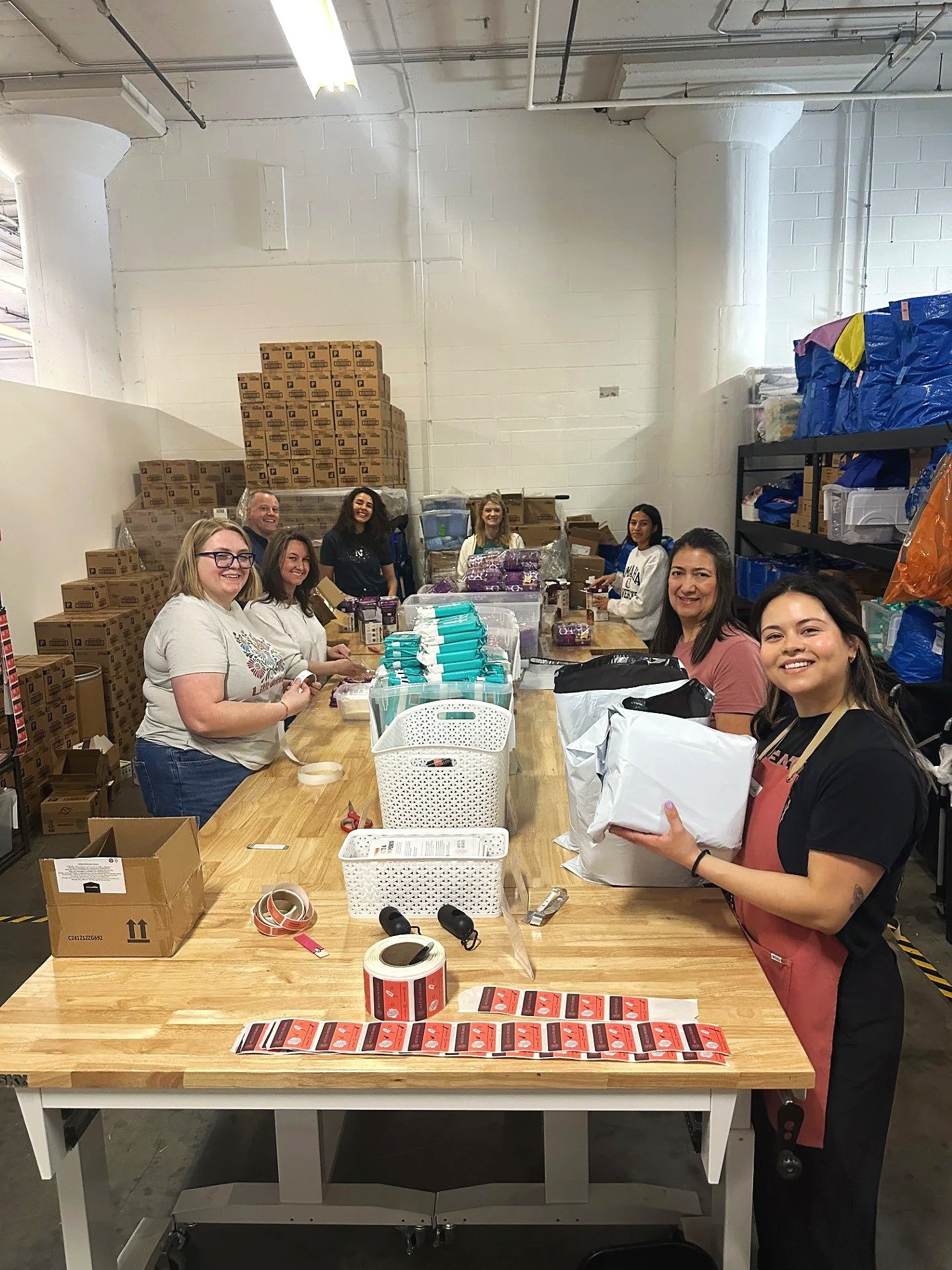 Group of people working together at a long table filled with supplies in a warehouse or storage room. There are boxes, packages, and packing materials on the table and shelves.