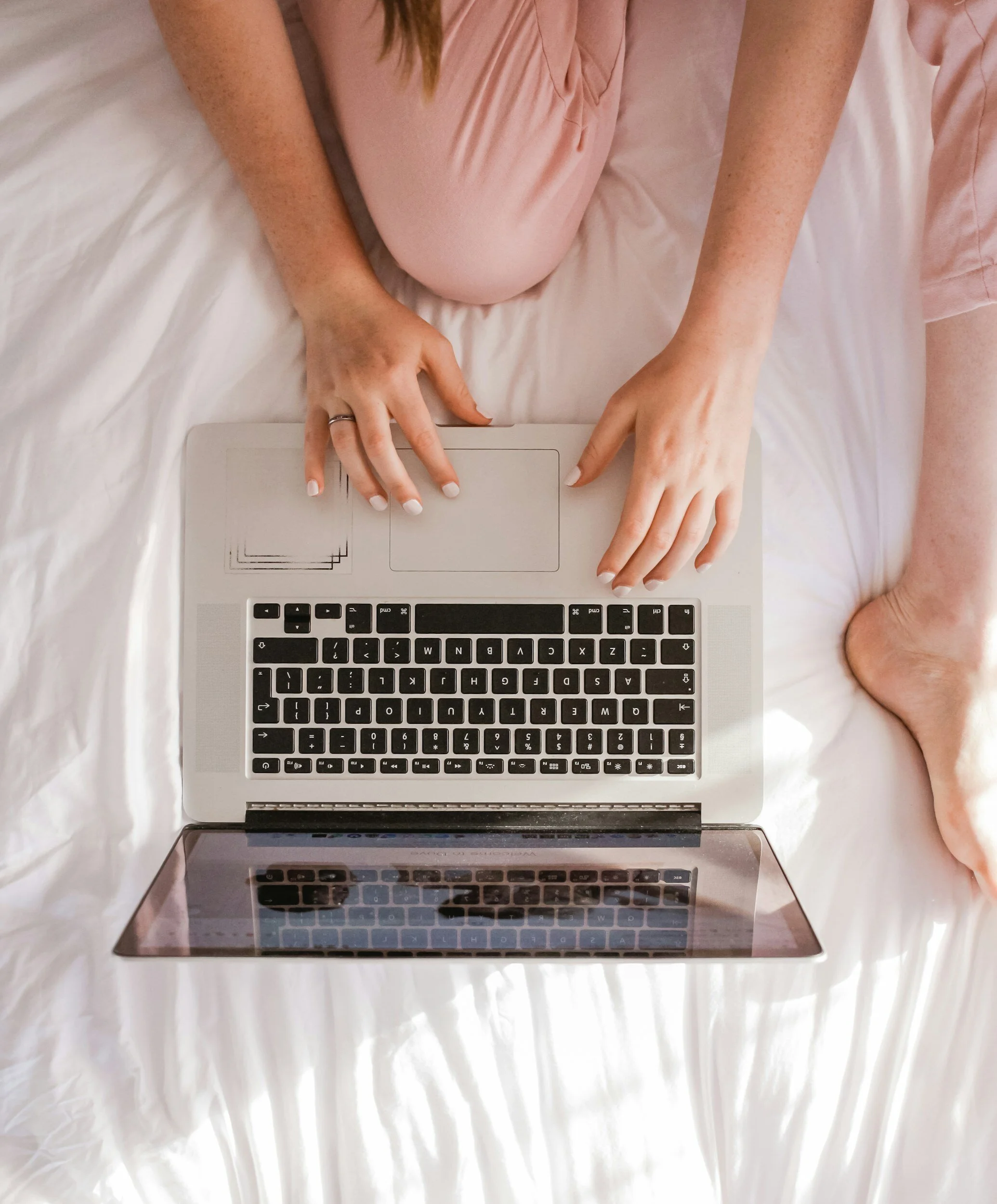 Top view of a person sitting on a bed with a white bedsheet, wearing a pink outfit, using a silver laptop with a black keyboard and a guide on the screen.