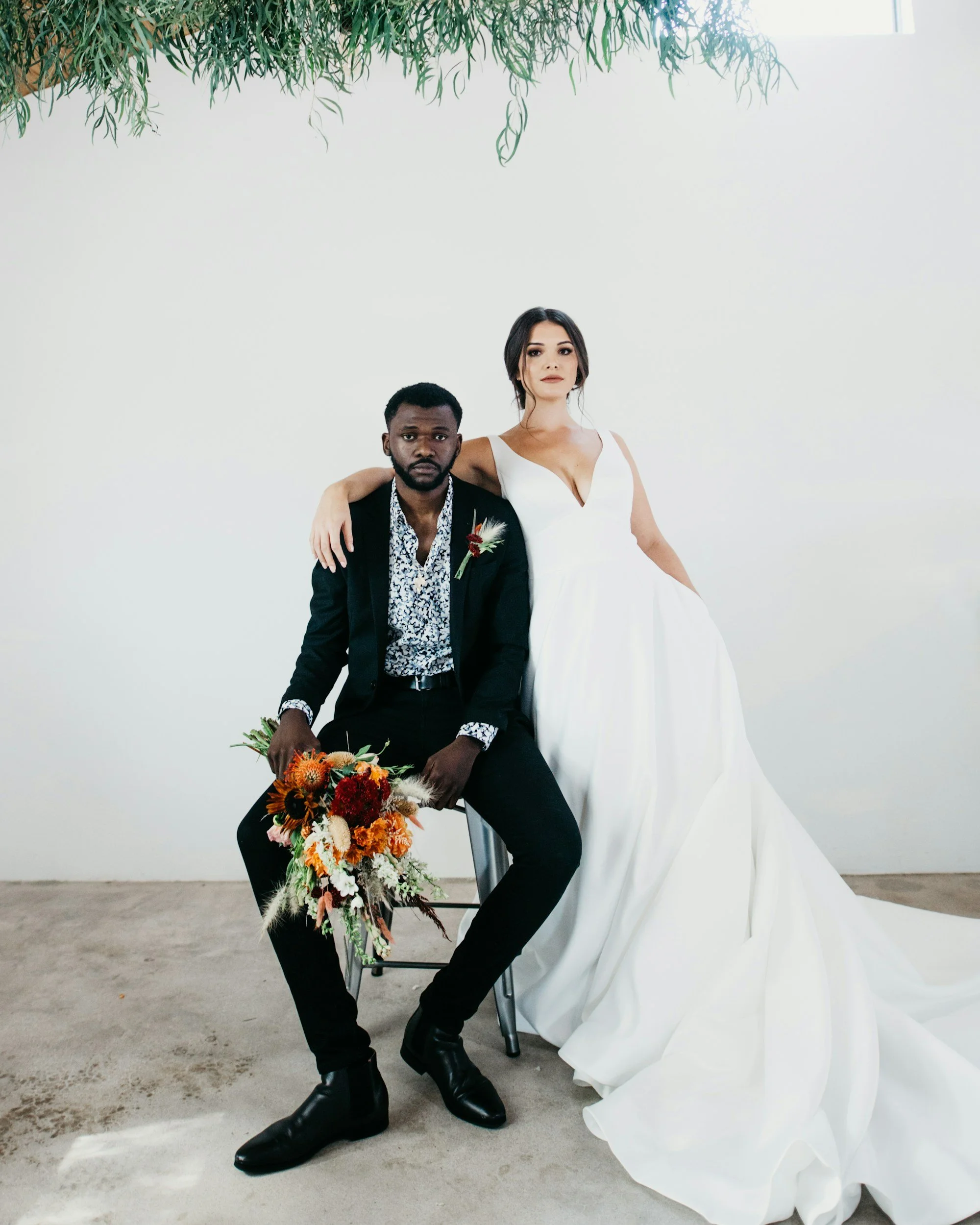 A man and a woman in wedding attire, with the woman in a white wedding dress and the man in a black suit, sit close together indoors against a plain white wall with greenery hanging from above, the man holding a bouquet of orange and red flowers.