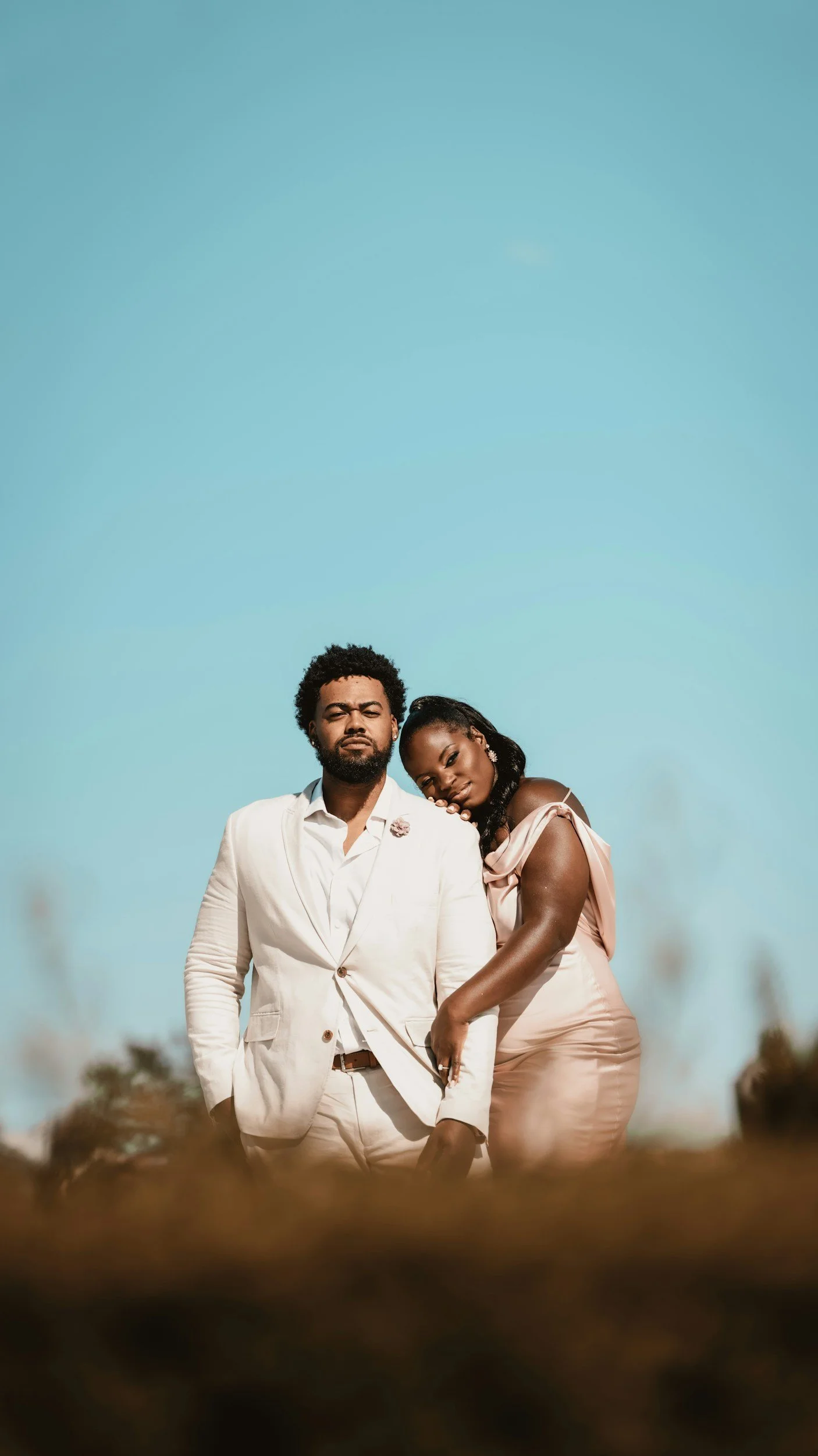A couple stands closely together outdoors under a clear blue sky, with the woman leaning her head on the man's shoulder. The man is wearing a white suit, and the woman is dressed in a blush pink dress.
