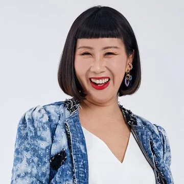Person with short black hair, wearing a blue patterned jacket and white blouse, smiling.