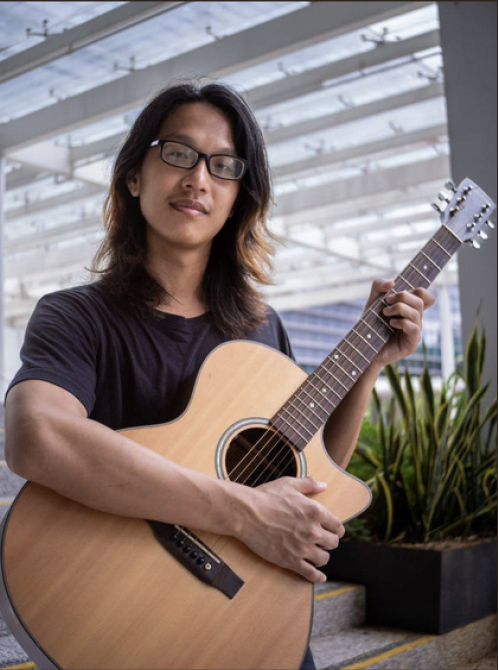 Person with long dark hair and glasses holding an acoustic guitar in an indoor space with glass ceiling and plants