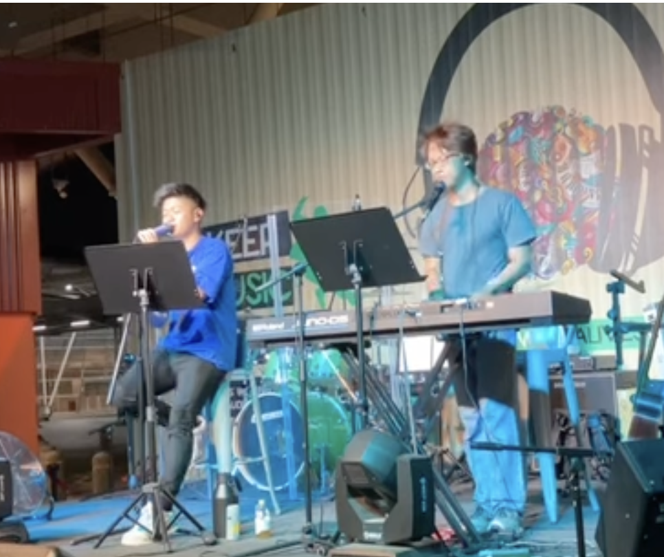 Two musicians perform on stage with one singing into a microphone and the other playing a keyboard. There are music stands and equipment around them.