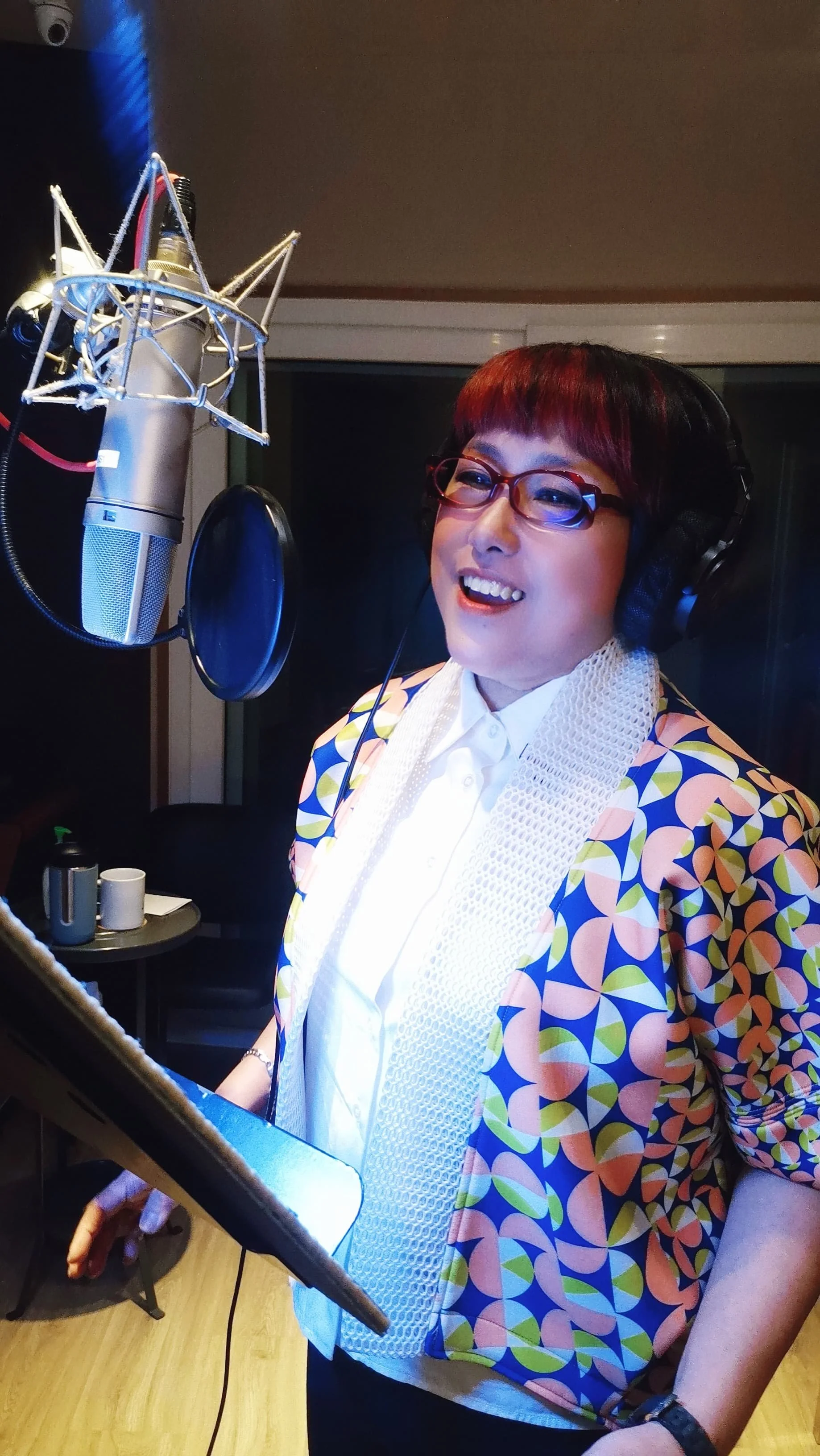 Person in recording studio with microphone and headphones, reading from a stand, wearing a colorful jacket.