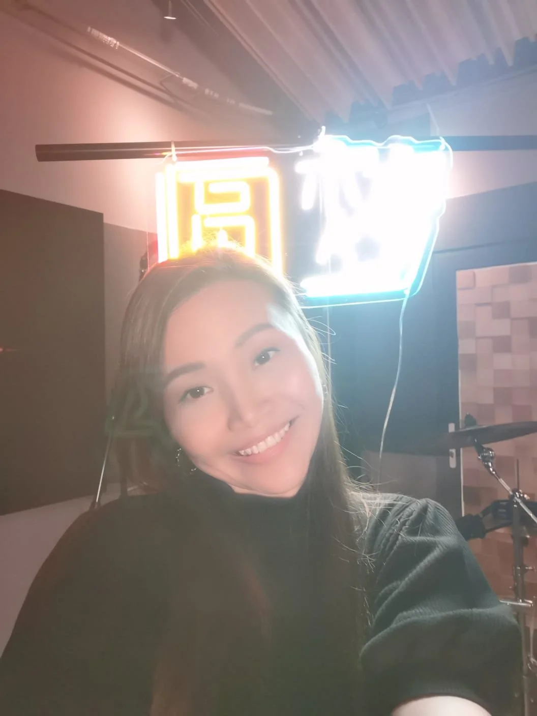 Person taking a selfie in front of a neon sign in a room with a drum set in the background.