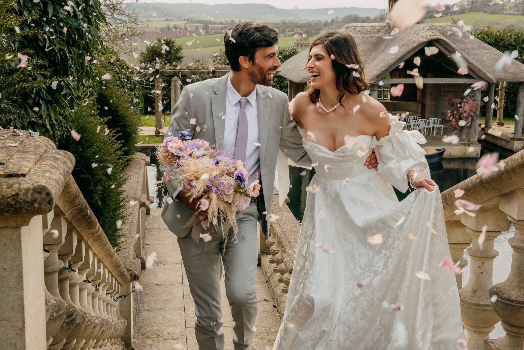 A bride and groom walking down an outdoor staircase, surrounded by falling flower petals. The bride is wearing a white dress, while the groom is in a light gray suit holding a bouquet. They are both smiling and appear happy, with a scenic background