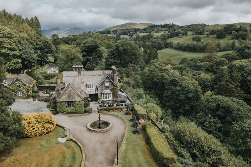 Aerial view of a large country house surrounded by lush green trees and landscape, with mountains in the background. A fountain is in the circular driveway in front of the house.