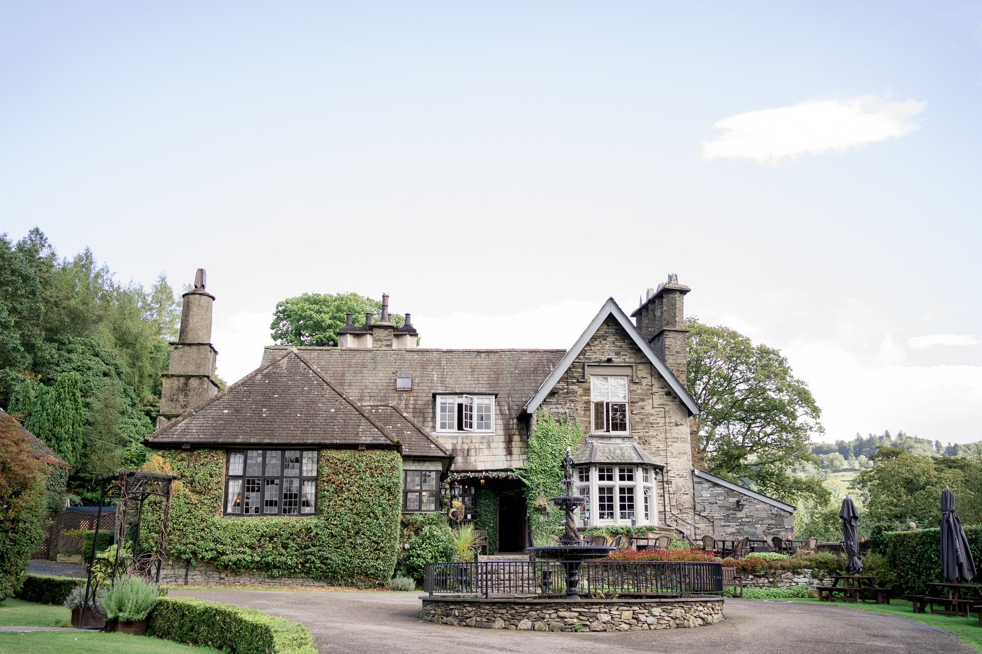 Stone country house with ivy-covered walls, surrounded by greenery, featuring a fountain and outdoor seating in the garden.