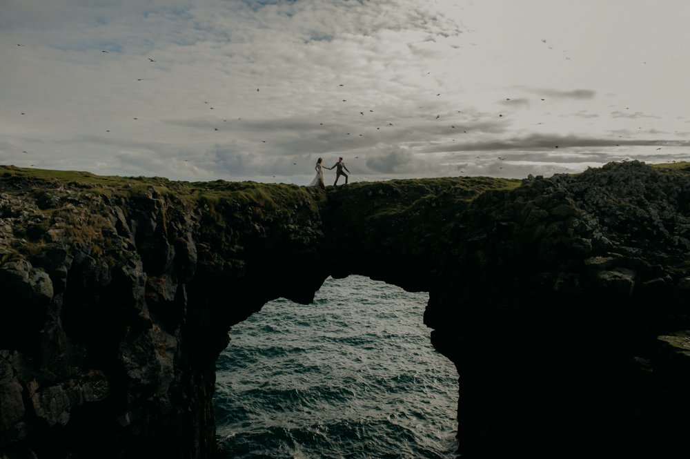 Two people standing on a rock arch above the ocean under a cloudy sky.
