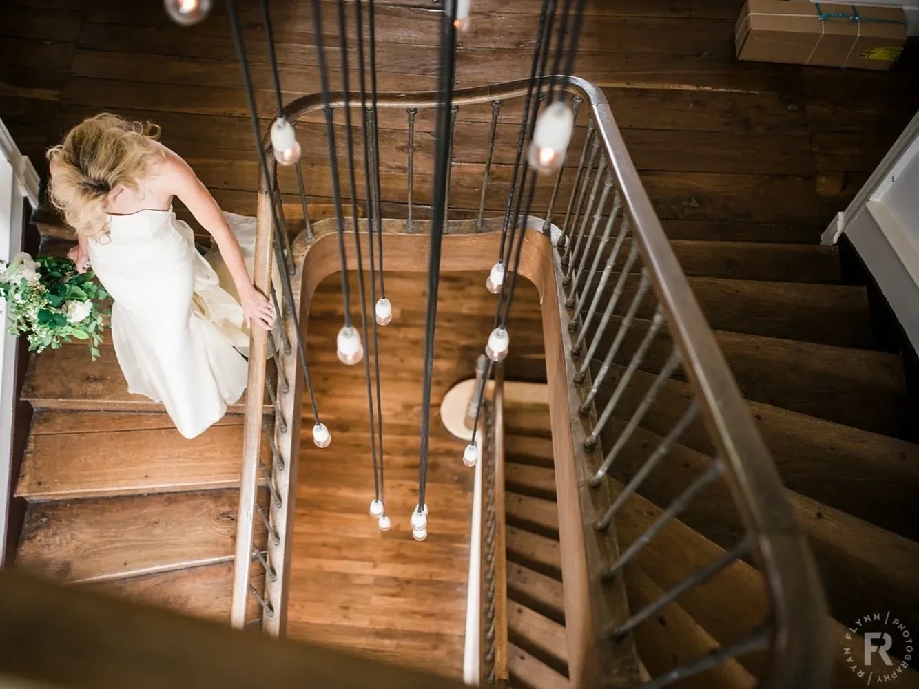 A woman in a white dress walking down a wooden staircase seen from above, with string lights hanging from the ceiling.